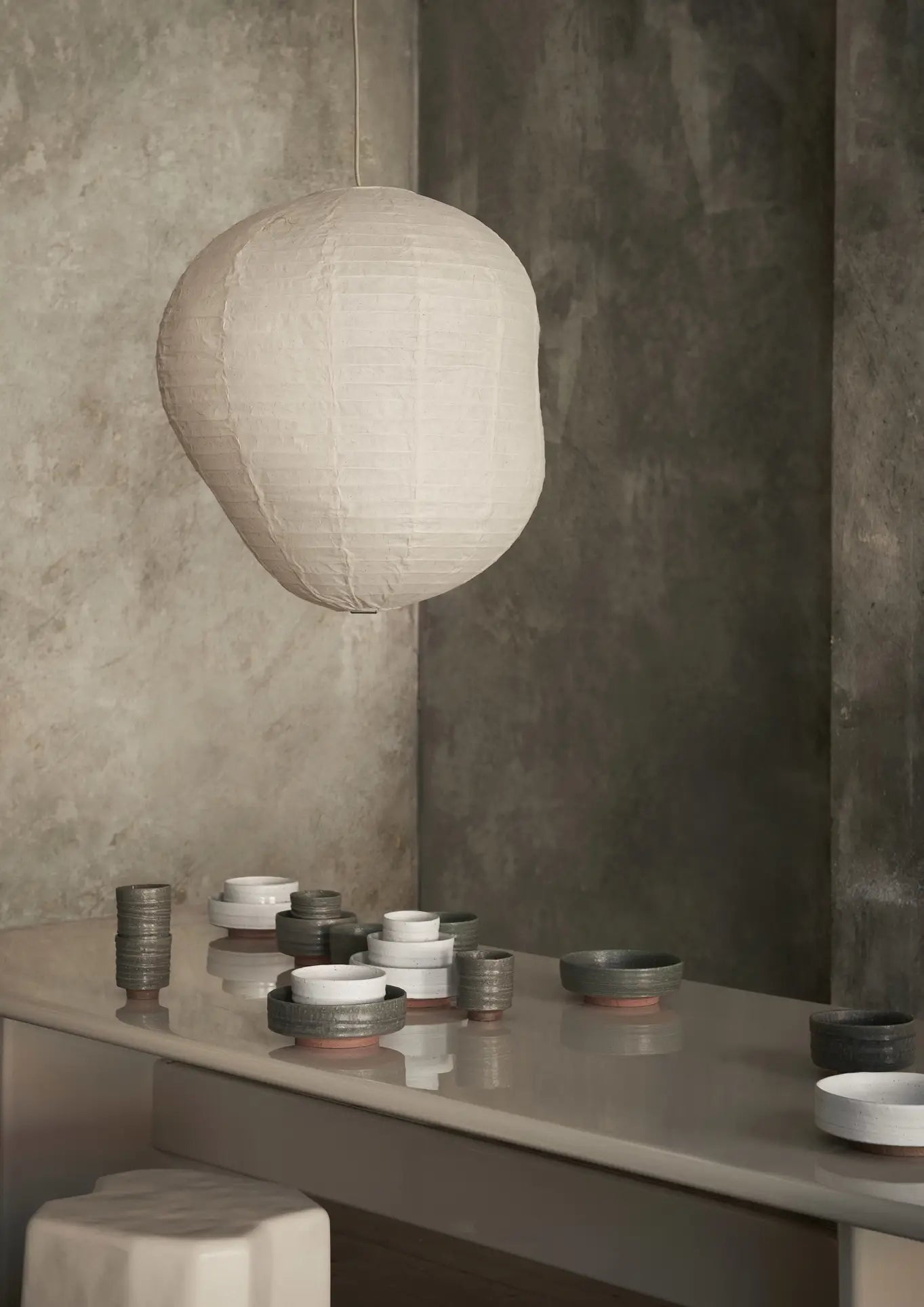A soft, white paper lantern hangs above a glossy table set with neatly arranged ferm LIVING Serena Bowls (12.5cm) and cups in neutral tones, against a textured gray wall in a modern, serene interior.