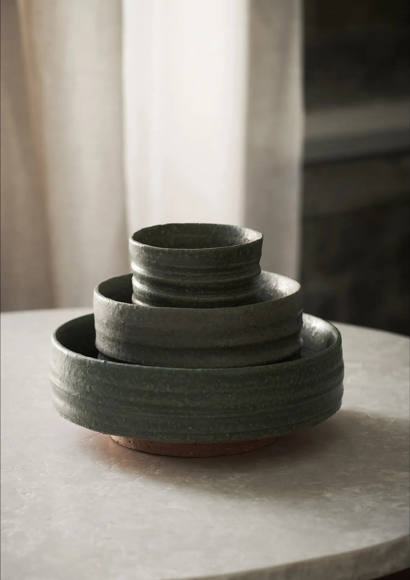 The ferm LIVING Serena Bowl (12.5cm) by ferm LIVING, in dark green ceramic with a luxurious texture, is stacked with matching bowls on a light round table, soft neutral curtains in the background.