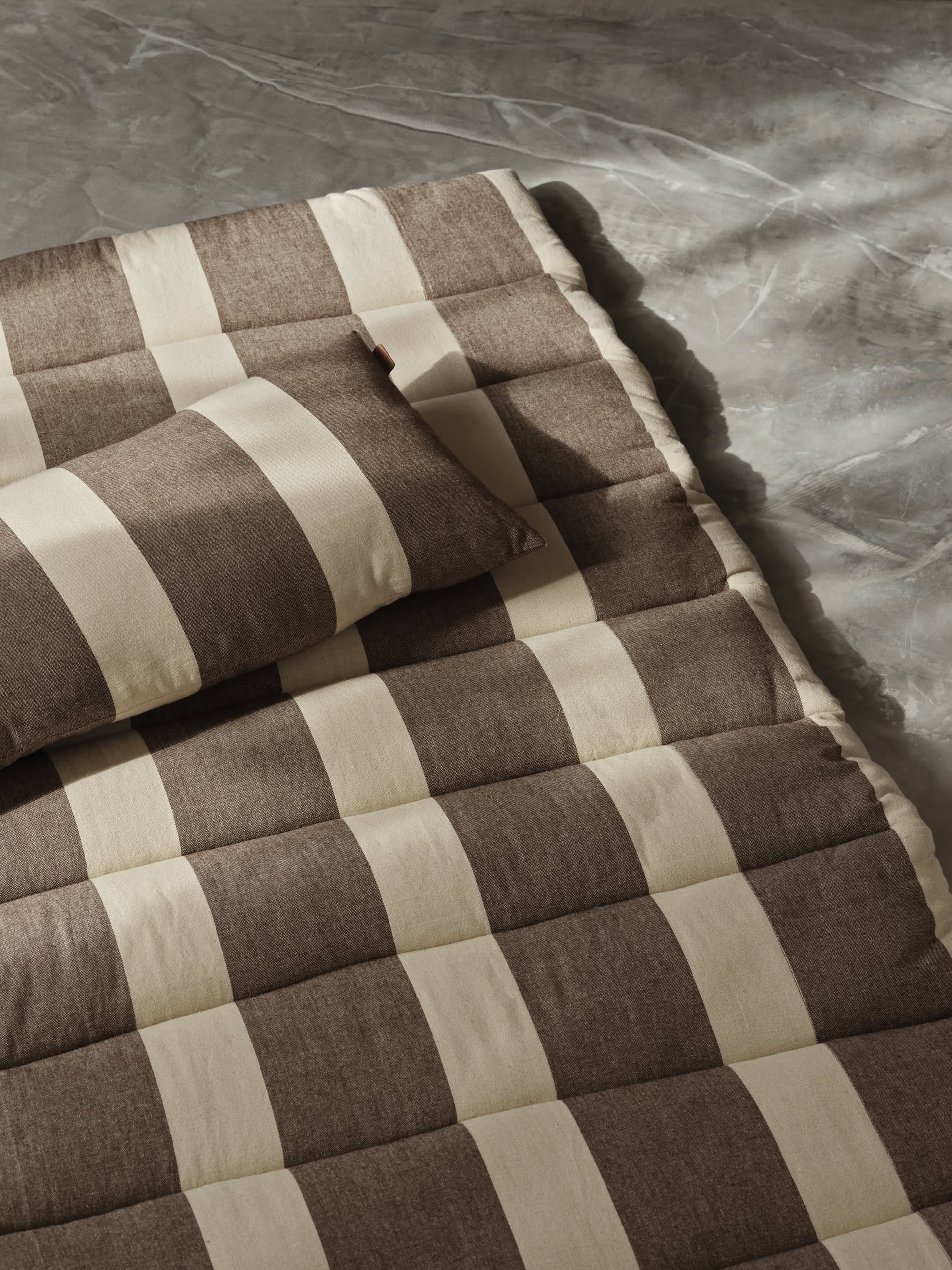 The ferm LIVING Strand Quilted Mattress, featuring a brown and beige checkered organic cotton design with a matching pillow, rests on a gray marble surface for a cozy, stylish look.