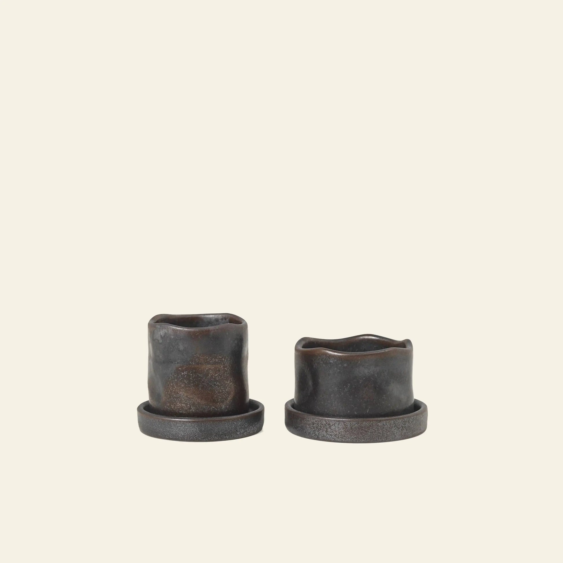 The ferm LIVING Uneru Mini Pots, Set of 2, features small dark stoneware pots with irregular rims and matching round saucers—ideal as a unique plant gift or for displaying your favorite ferm LIVING stoneware.