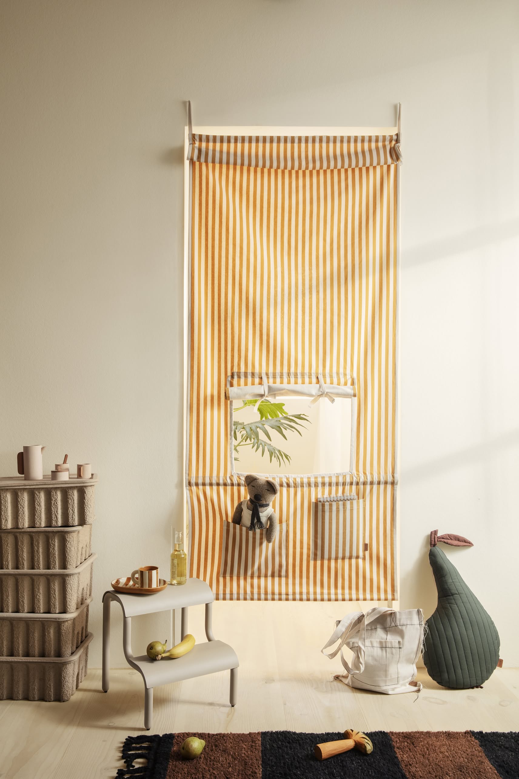A mustard yellow and white striped curtain with a small window hangs on a textured surface. A plush toy peeks out as shelves display cups, a banana, and decor—beneath it sits the ferm LIVING Up Step Stool by ferm LIVING.