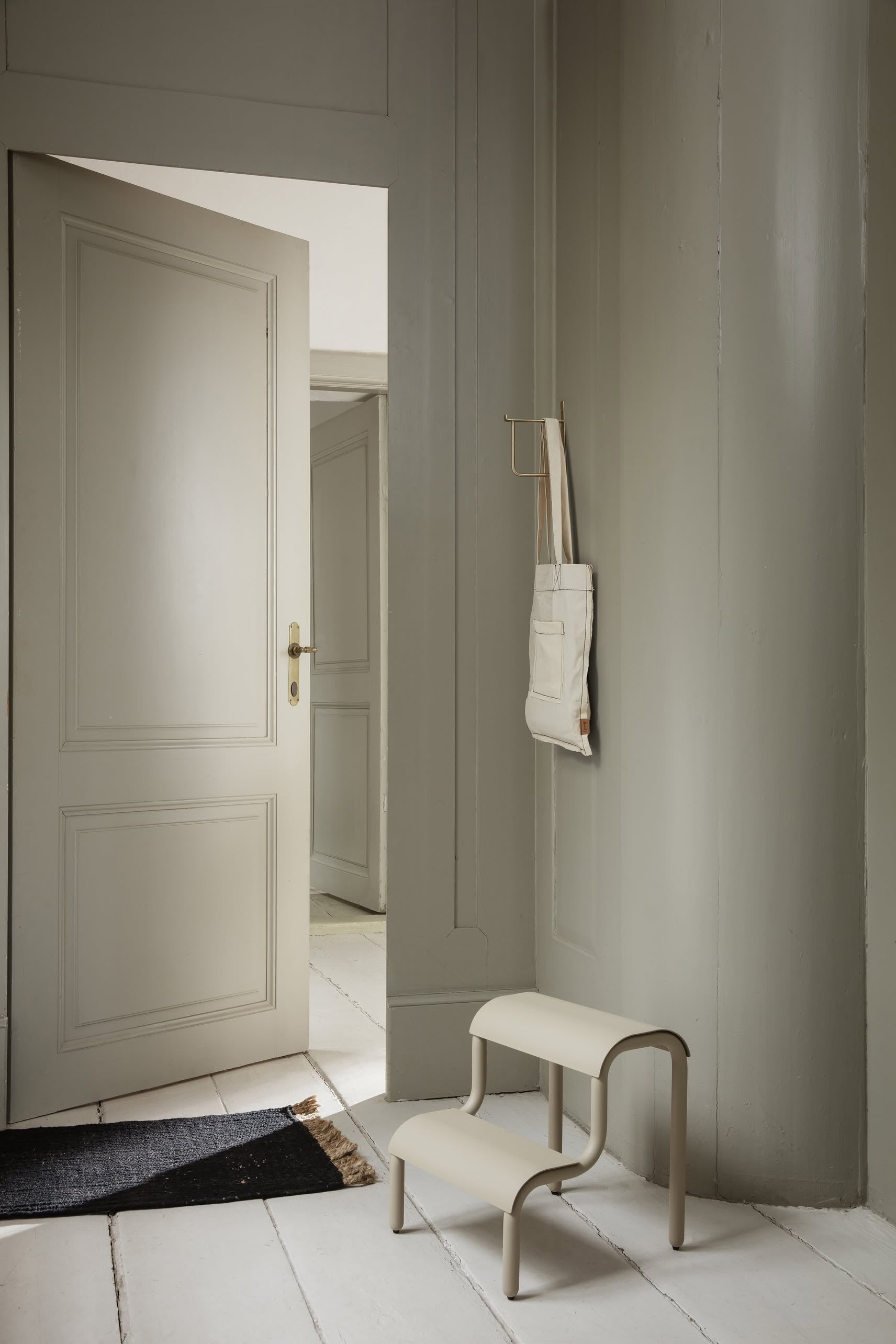 Minimalist hallway with light gray paneled walls, an open door, white wooden floor, the ferm LIVING Up Step Stool in beige with a textured surface, a small wall hook with a canvas tote bag, and a dark doormat by the entrance.