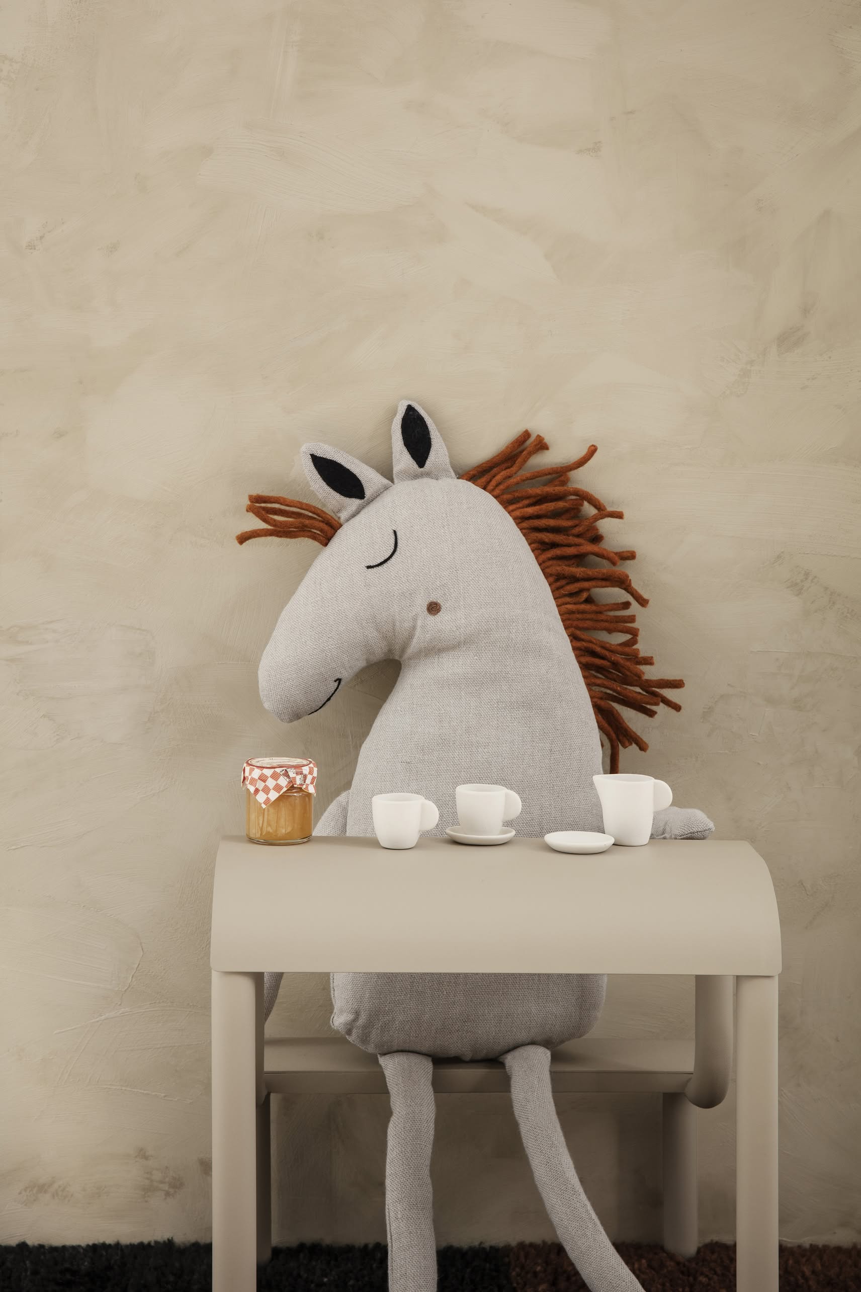 A large stuffed horse toy with closed eyes and a brown yarn mane sits at a small beige table with white toy teacups and honey beside a ferm LIVING Up Step Stool, all against a neutral wall background.