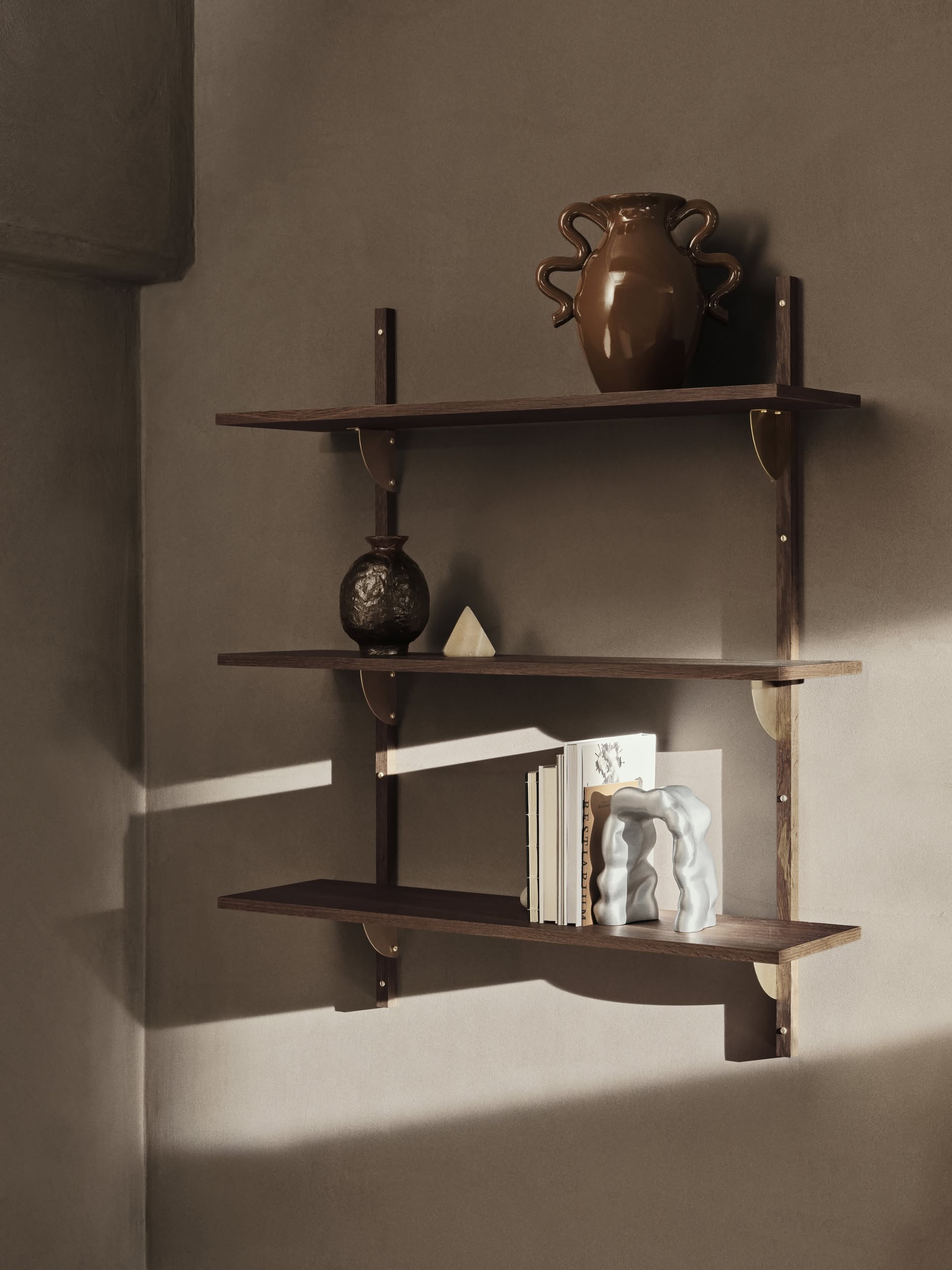 Three dark wooden shelves on a beige wall display a brown vase, small round pottery, the ferm LIVING Verso Table Vase with its organic silhouette, several standing books, and a geometric object. Warm light gives the scene a contemporary touch.