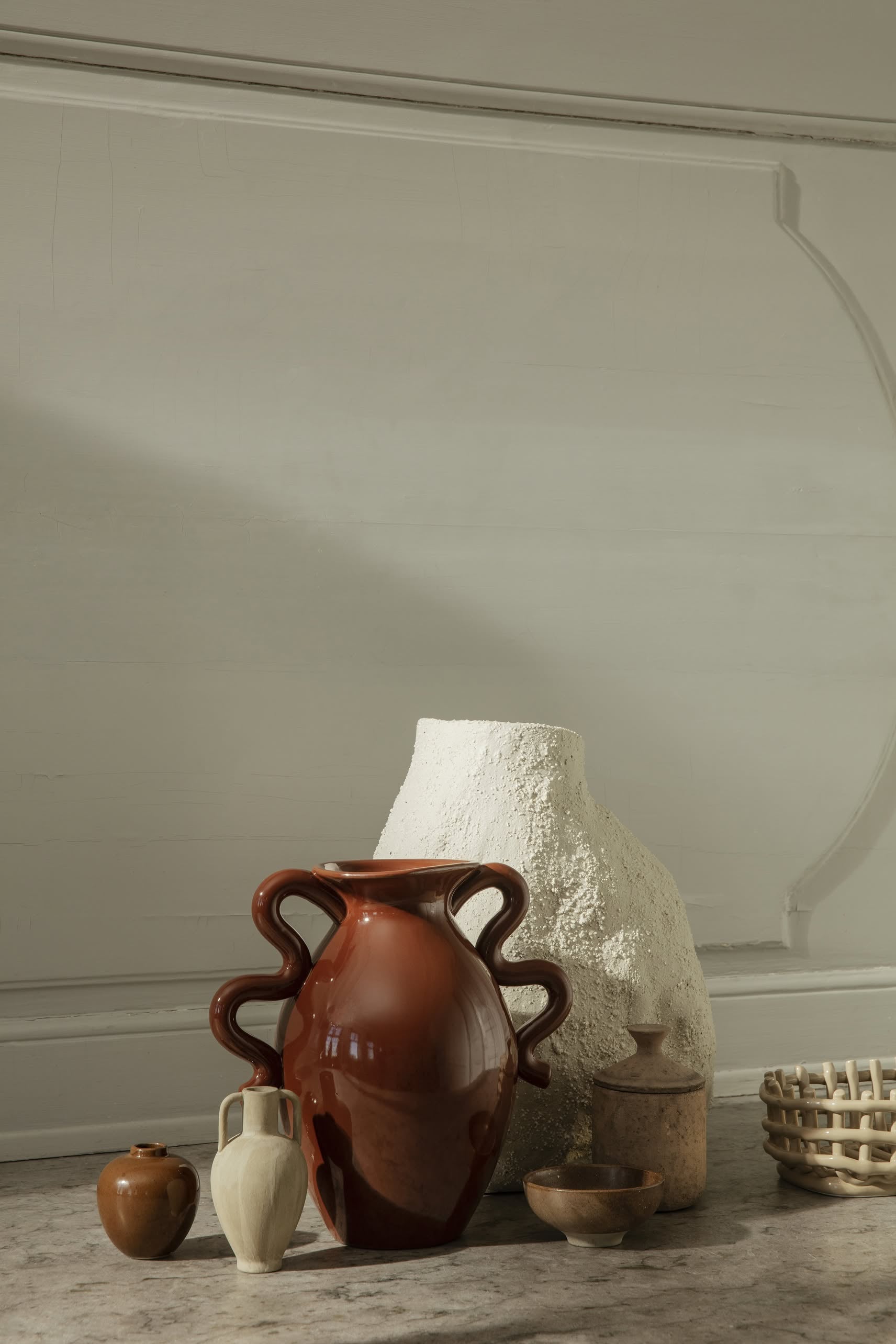 A contemporary still life features the ferm LIVING Verso Table Vase with its organic silhouette, a textured white vase, and small ceramics, all set against a light gray minimalist wall for modern appeal.