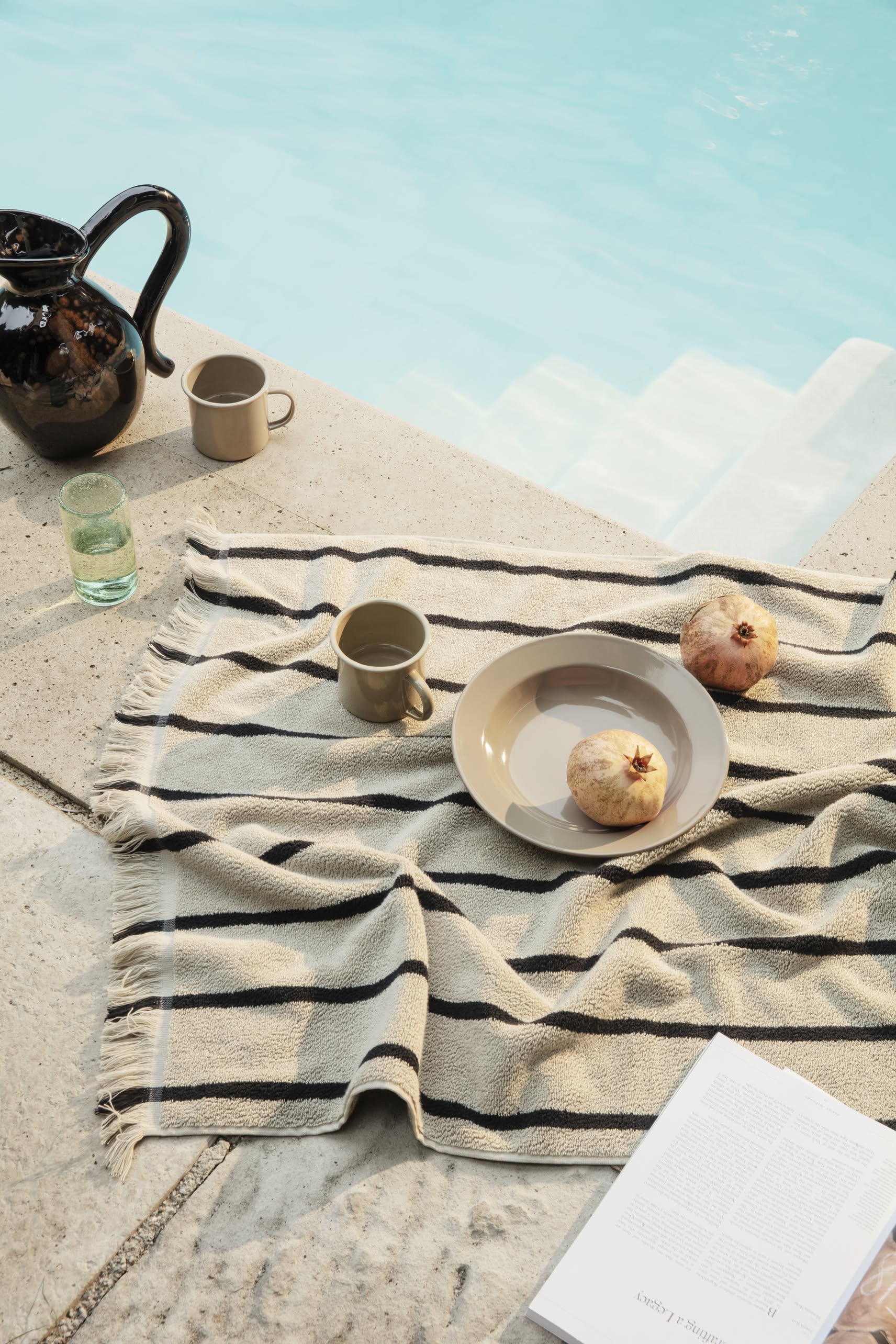 A striped beige and black towel is spread by the pool with the ferm LIVING Yard Picnic, Set of 8, featuring two pomegranates, a mug, a glass of water, a dark ceramic pitcher, and an open book arranged for relaxed outdoor dining.