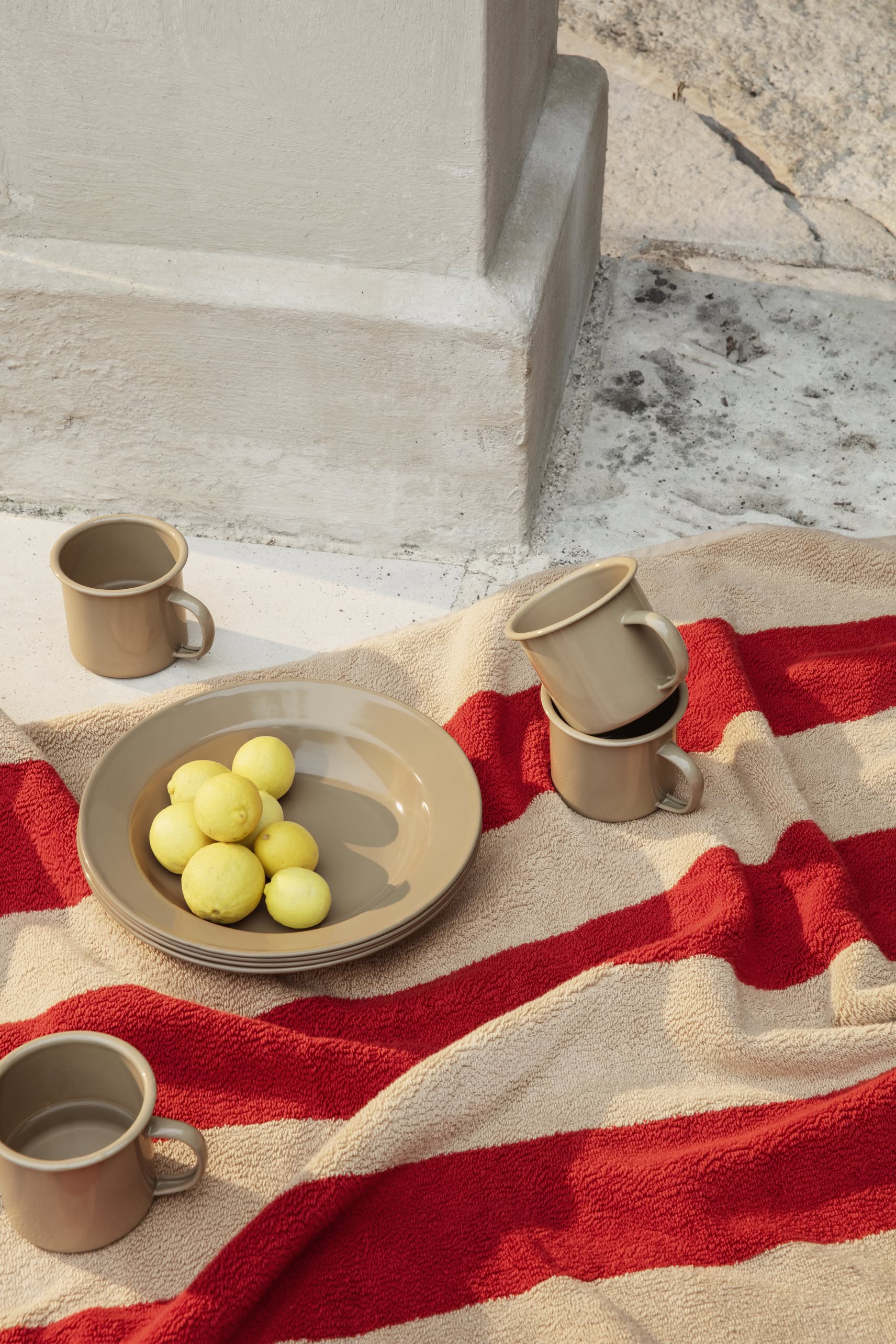 A ferm LIVING Yard Picnic, Set of 8 is displayed on a stone surface outdoors, featuring striped textiles, enamelware mugs, a beige plate with yellow fruit, and stacked mugs near a stone structure.