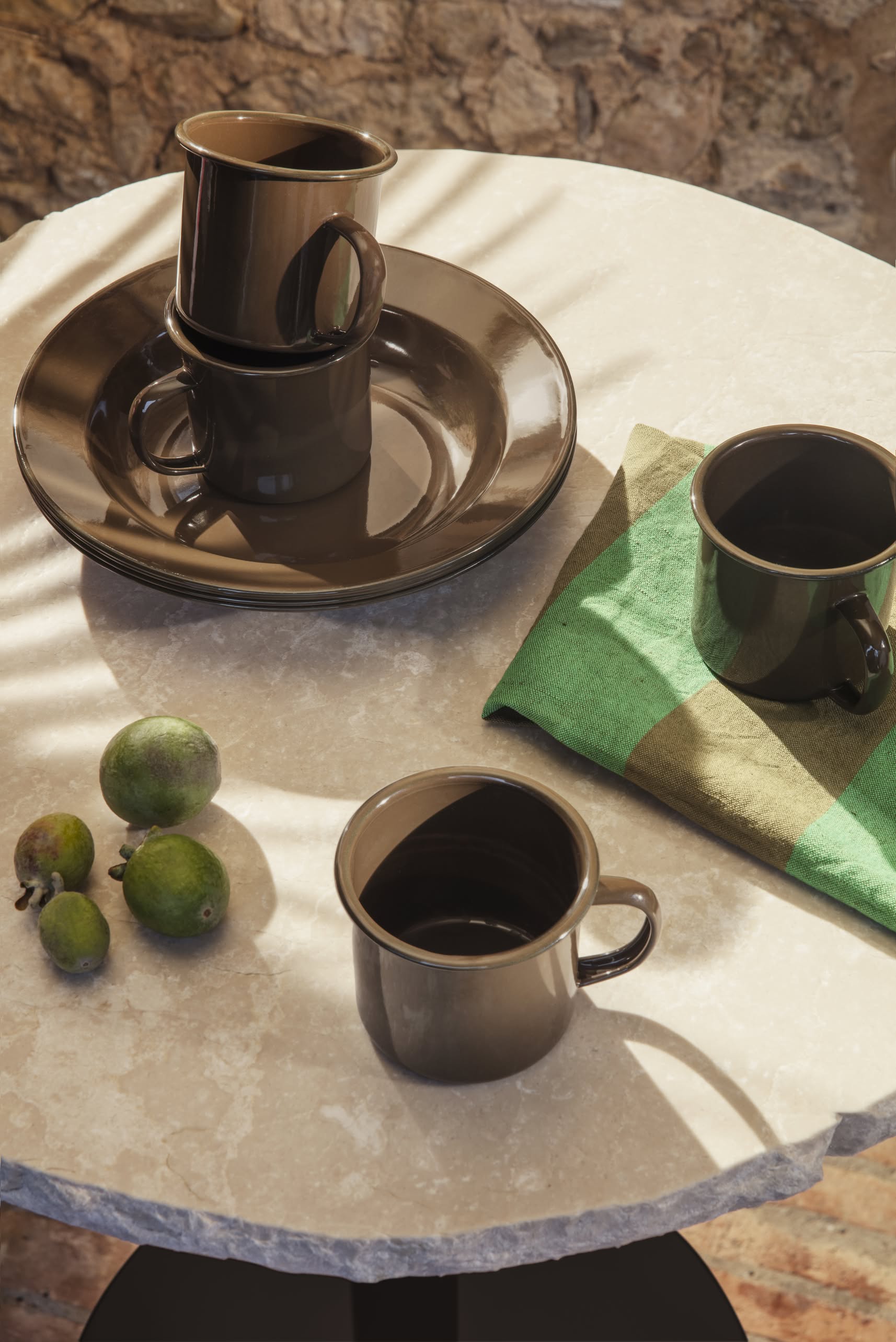 The ferm LIVING Yard Picnic, Set of 8 by ferm LIVING features dark brown cups and plates, a green checkered napkin, and small green fruits atop a round stone table, bathed in sunlight with leaf shadows for an inviting outdoor scene.