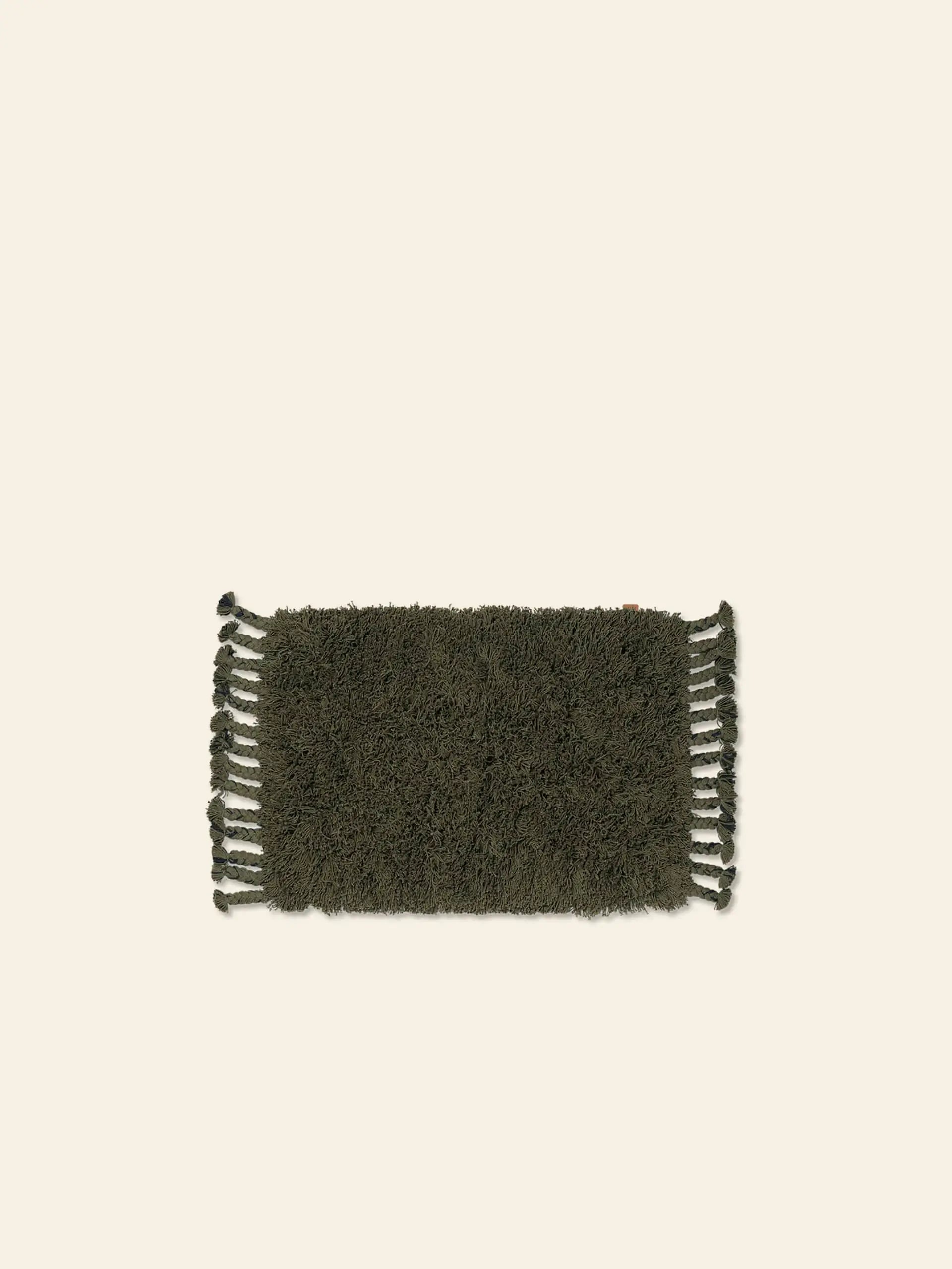 The ferm LIVING Amass Long Pile Mat is a rectangular, dark green shaggy rug with fringes on the short edges, shown on a light beige background.