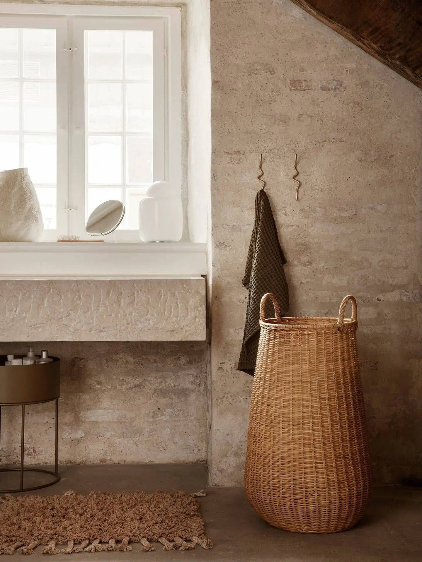 A cozy, minimalist room features the ferm LIVING Amass Long Pile Mat, a large woven basket, towels on hooks, and a white windowsill with small decor—set against a rustic stone wall and bathed in natural light.