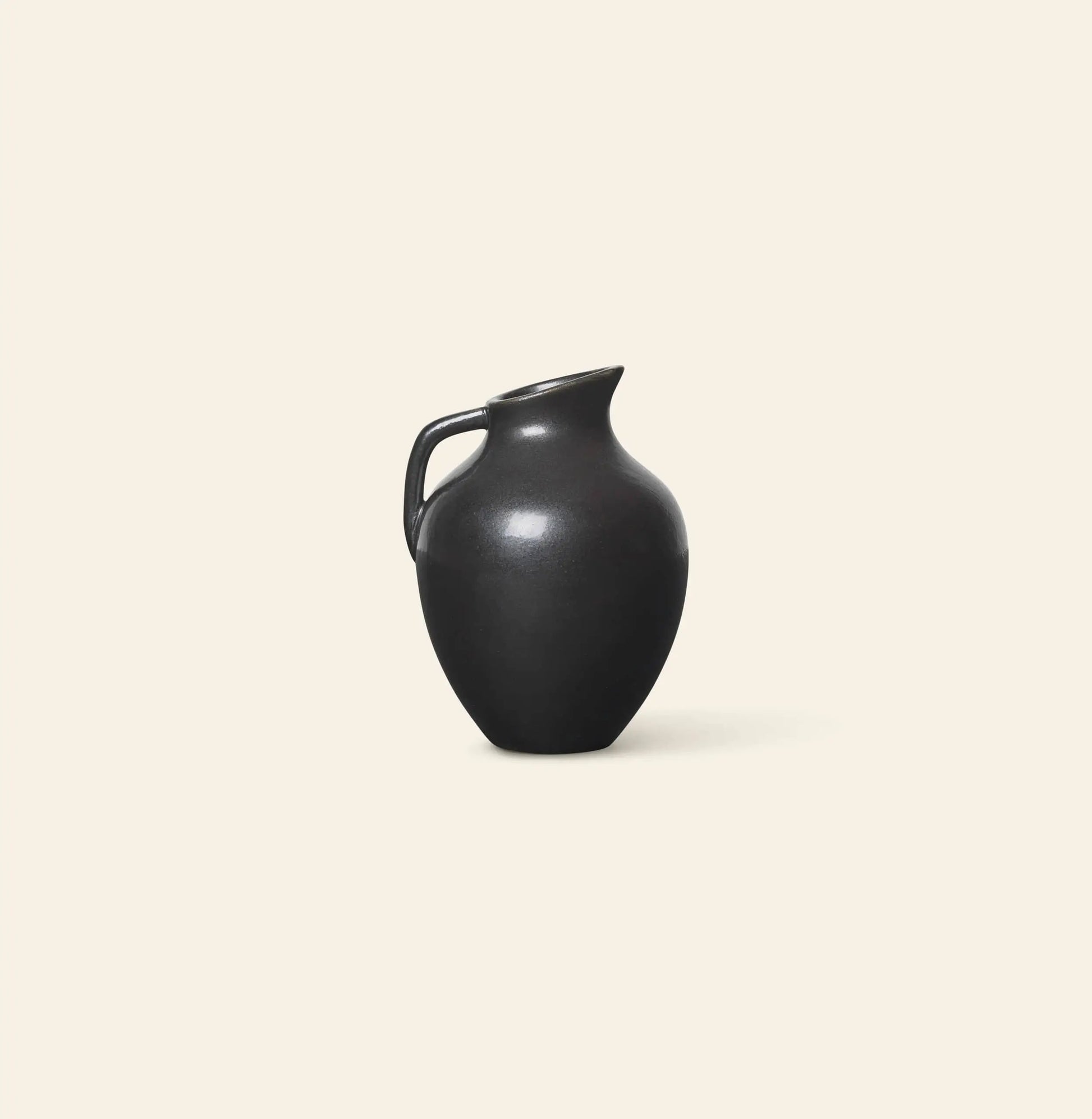 The ferm LIVING Ary Mini Vase M, by ferm LIVING, features a smooth black ceramic body with a curved handle and narrow spout. Its simple, elegant design echoes the timeless beauty of ancient Greek vases against a light beige background.