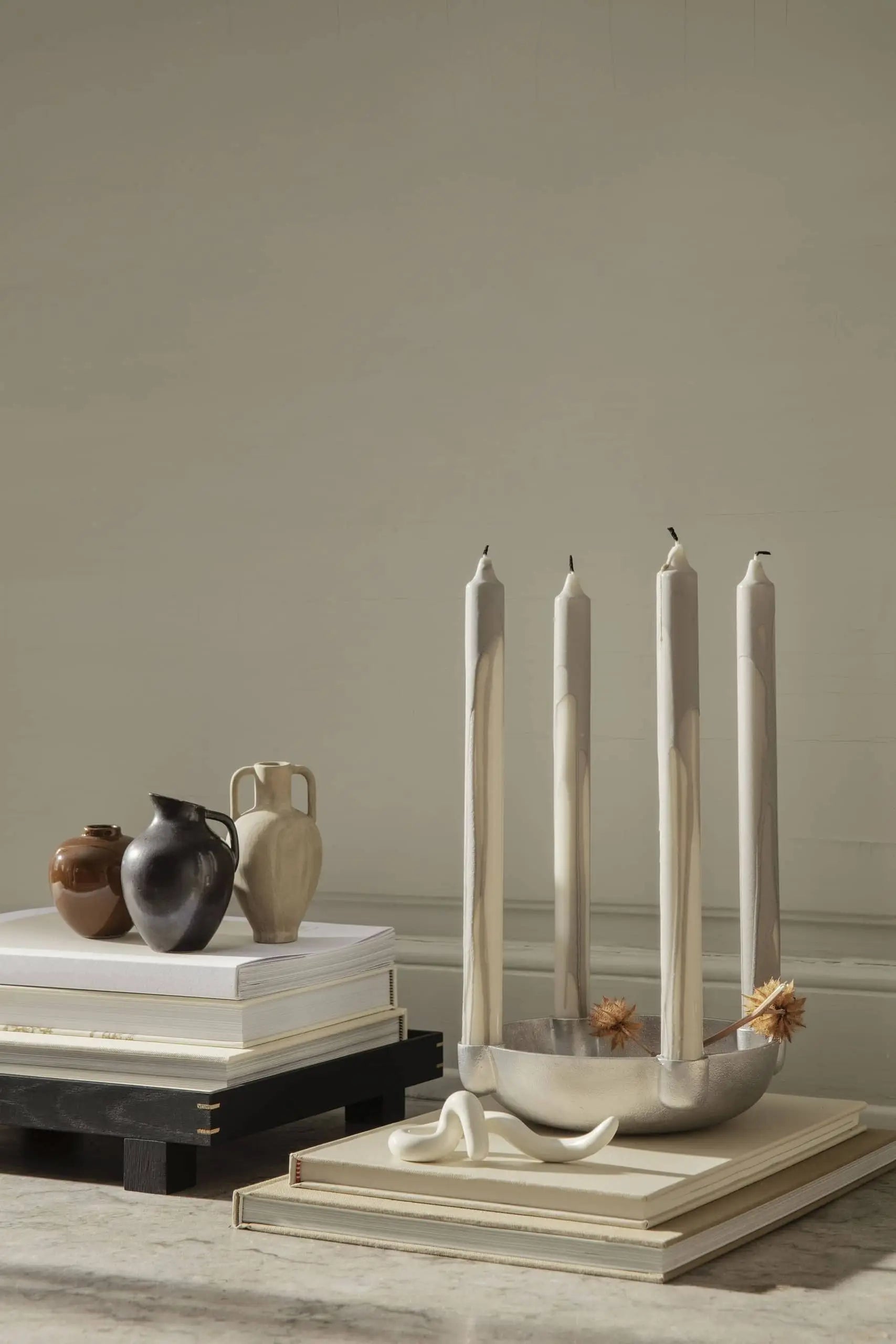 A minimalist display with four beige marbled candles in a silver holder, flanked by the ferm LIVING Ary Mini Vase M and small brown ceramics on stacked books, all set against a neutral beige wall.