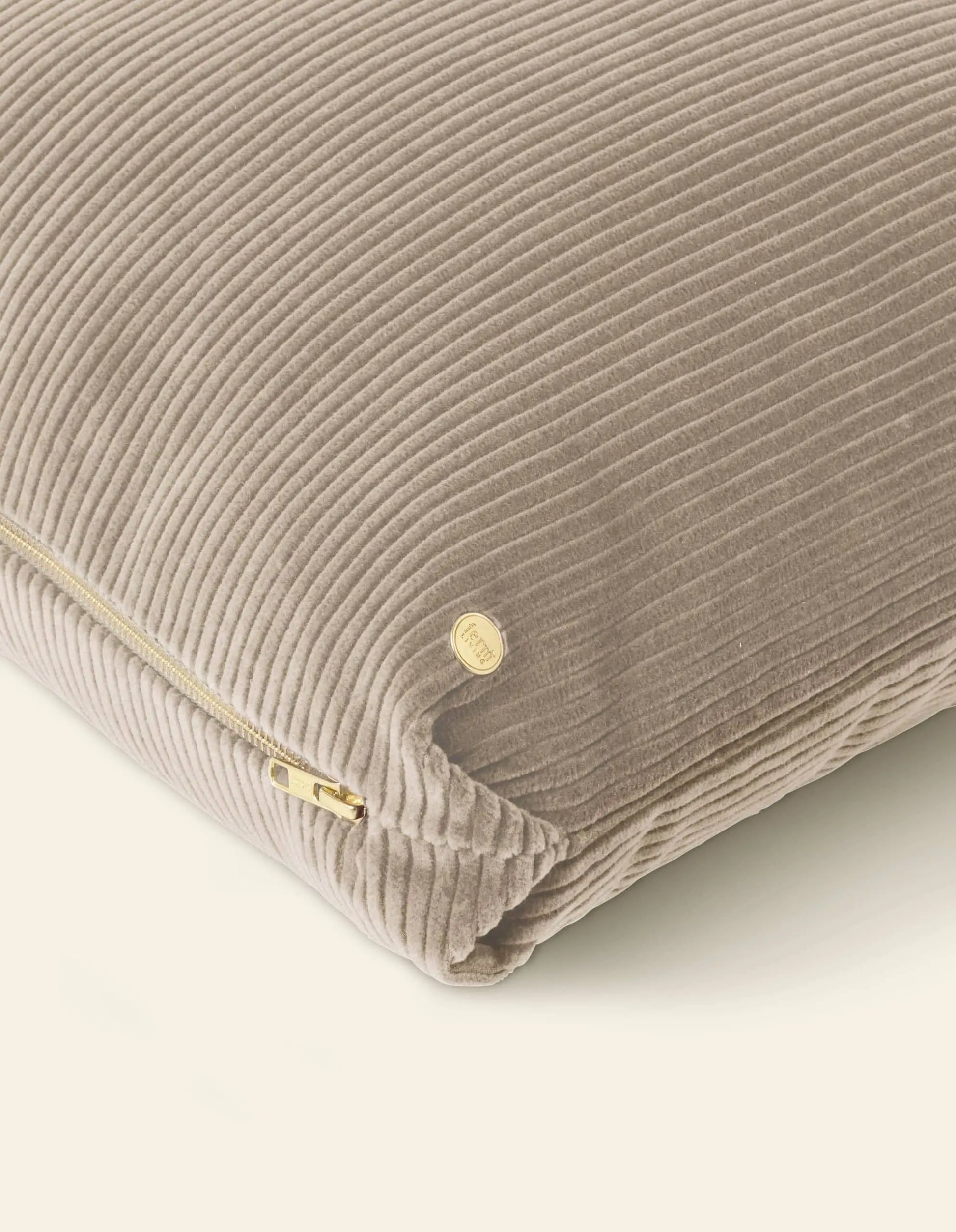 A close-up of the ferm LIVING Corduroy Cushion in beige with a corduroy texture, gold zipper, and a small round gold tag, set against a light background.