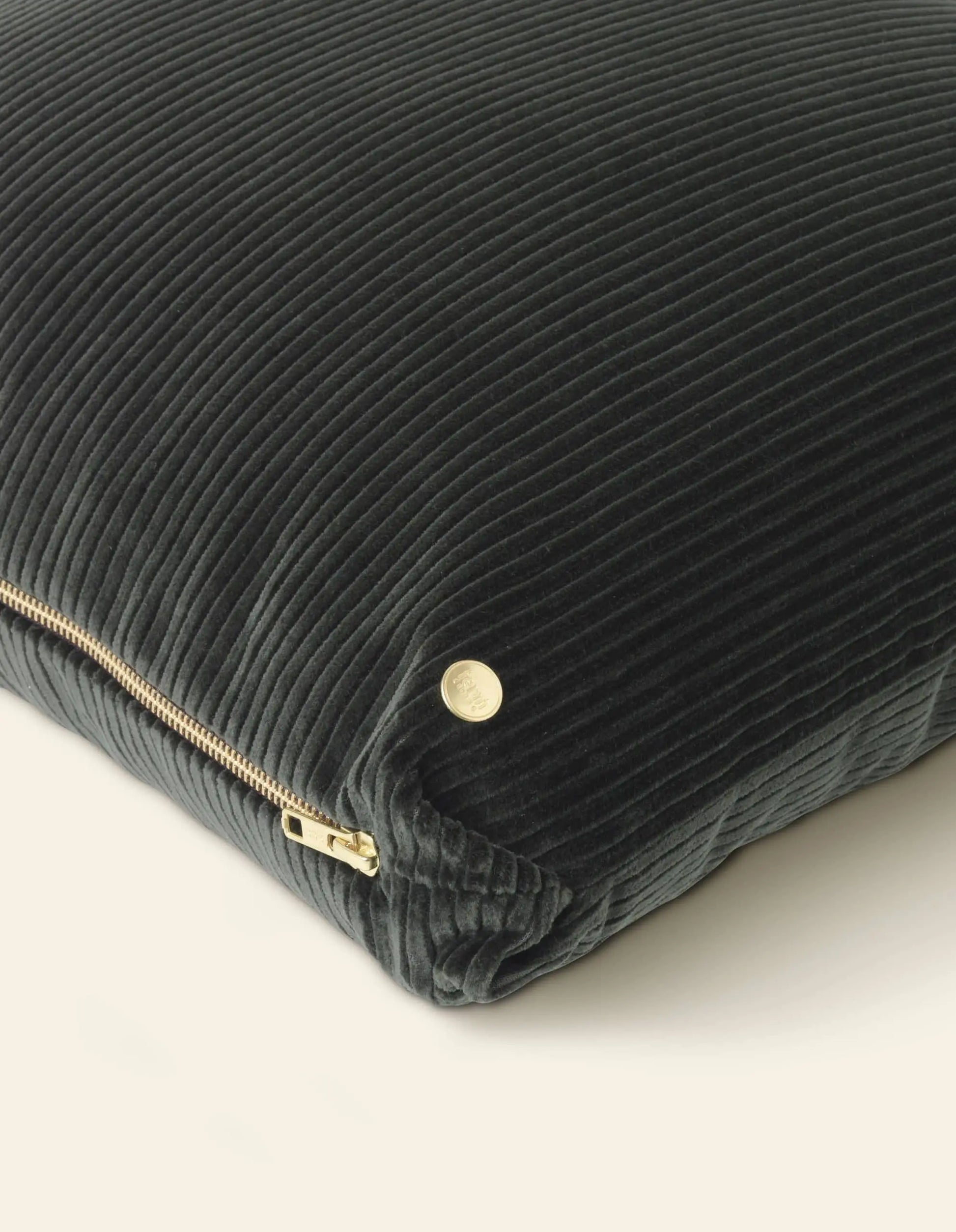 A close-up of the ferm LIVING Corduroy Cushion showcases its dark fabric with a visible gold zipper and a round gold tag on the corner, all set against a light background.