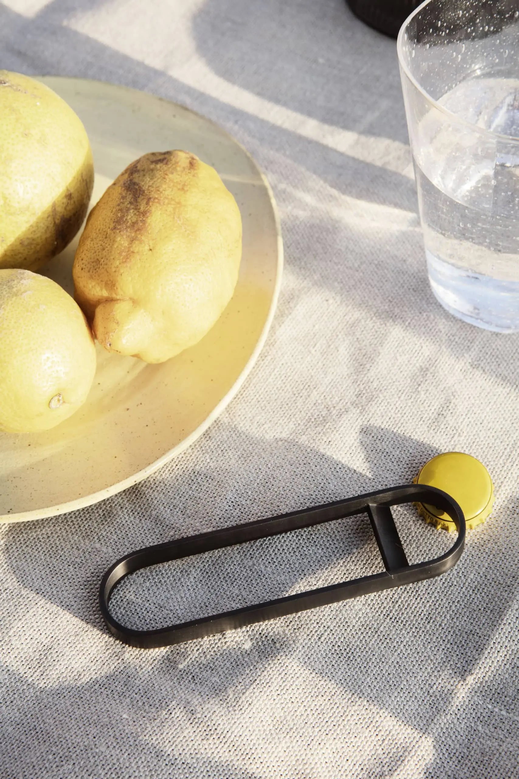 A plate with three lemons rests on a textured beige cloth in a modern kitchen, accompanied by a glass of water and the sleek ferm LIVING Fein Bottle Opener. Sunlight casts soft shadows across the minimalist space.