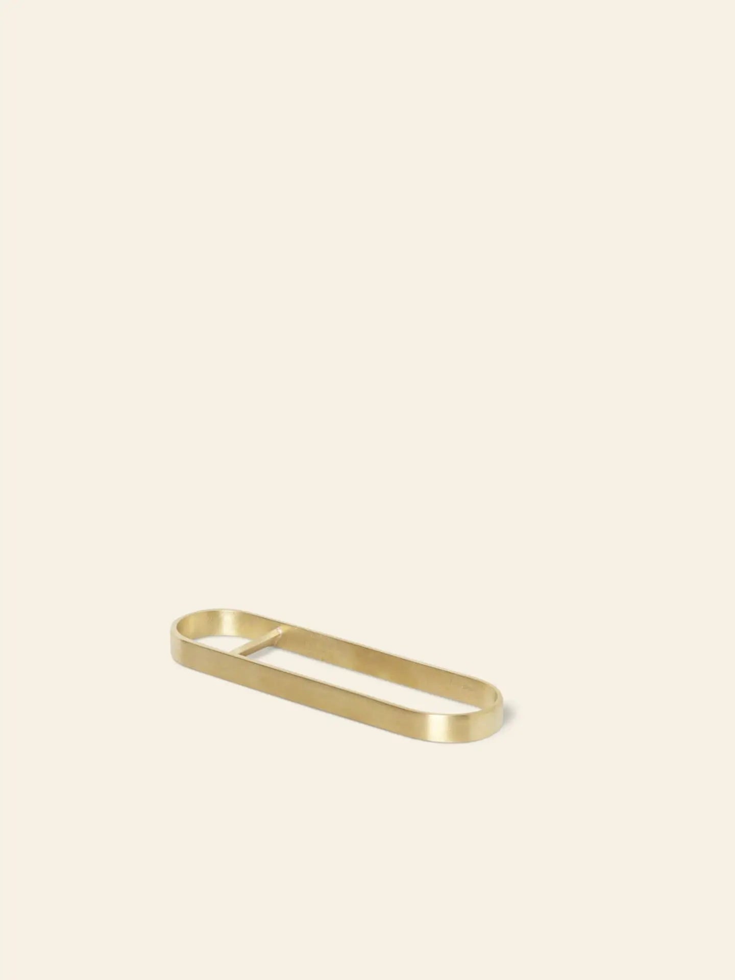The ferm LIVING Fein Bottle Opener, a minimalist gold metal accessory with an elegant oval design and central divider, adds a contemporary touch to any kitchen. Displayed on a simple light beige background.
