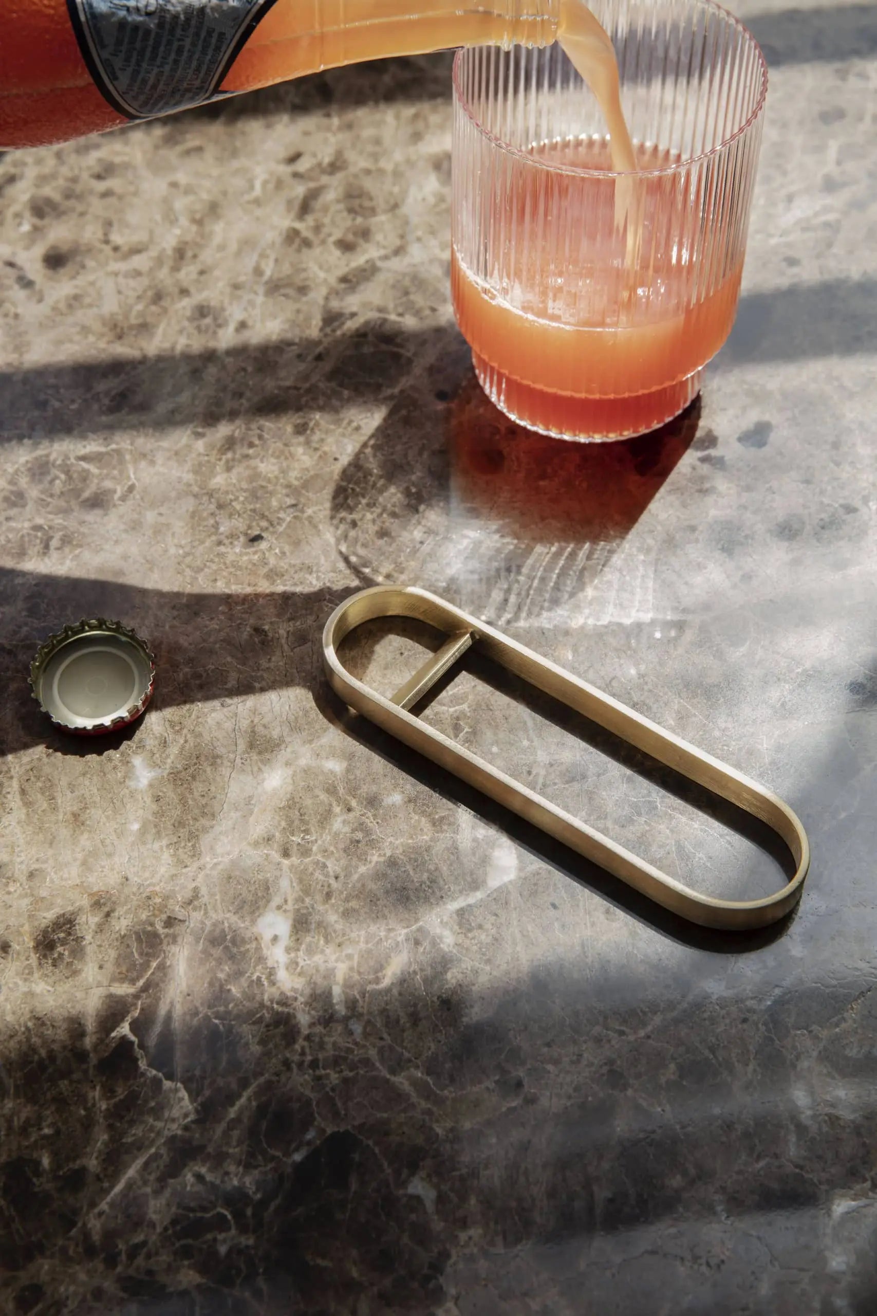 A glass of pinkish-orange drink sits on a marble table in a modern kitchen, next to the brass ferm LIVING Fein Bottle Opener and a green bottle cap. Sunlight casts soft shadows, emphasizing the minimalist style.