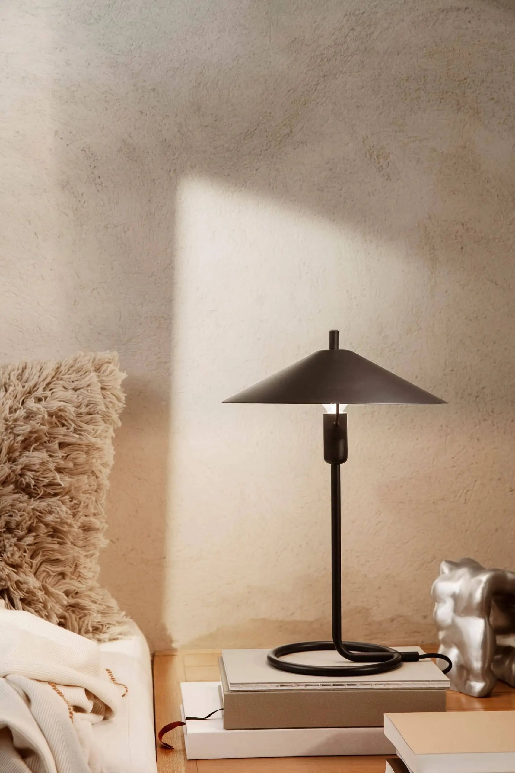 The ferm LIVING Filo Table Lamp, featuring a cone-shaped, powder-coated iron shade, sits on books beside a silver accent and textured beige pillow as sunlight casts geometric shadows—ideal for a modern home’s cream wall.
