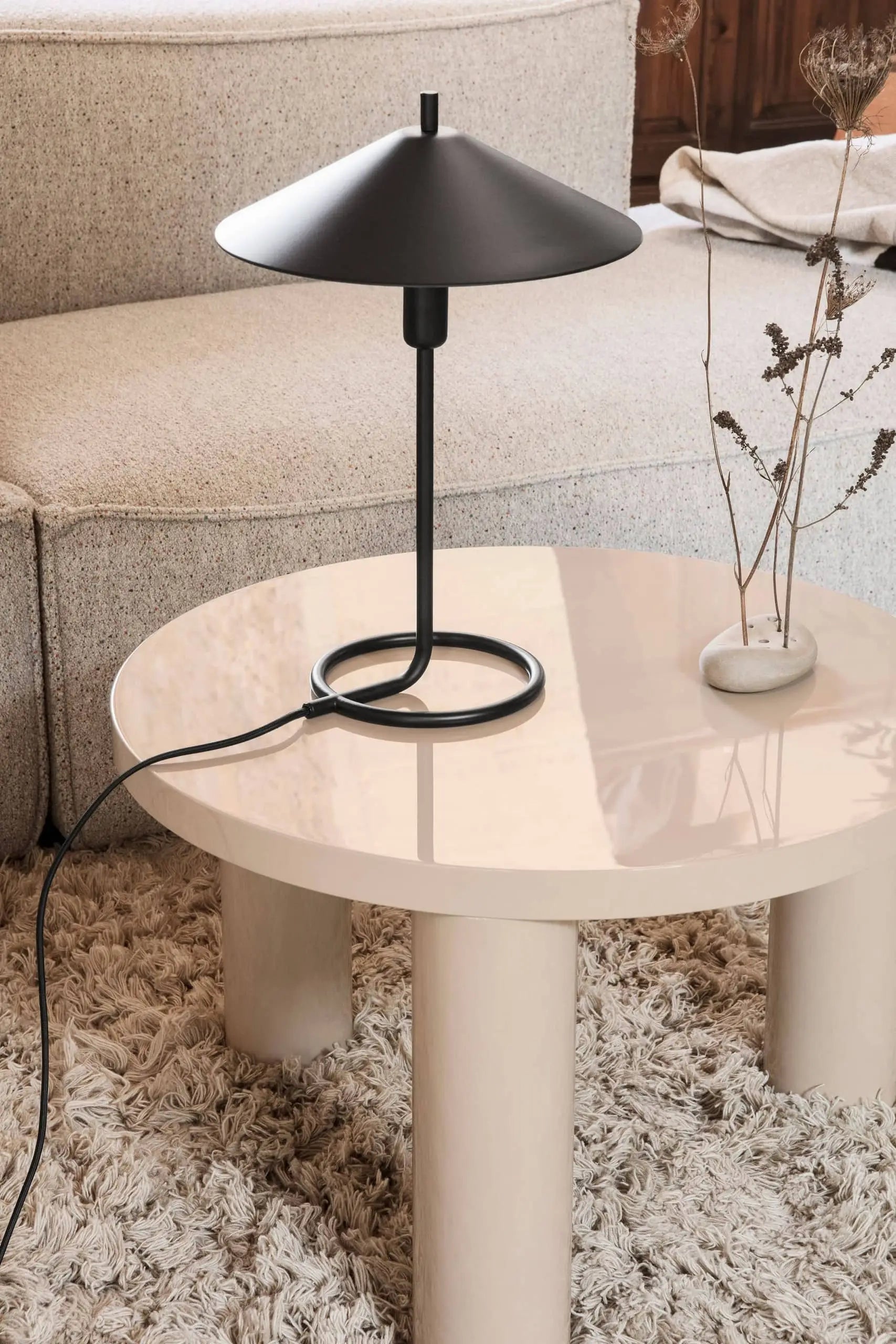 The ferm LIVING Filo Table Lamp, featuring a round shade and powder-coated iron base, is displayed on a glossy beige coffee table in a modern home with a textured rug, beige sofa, and a vase of dried branches.