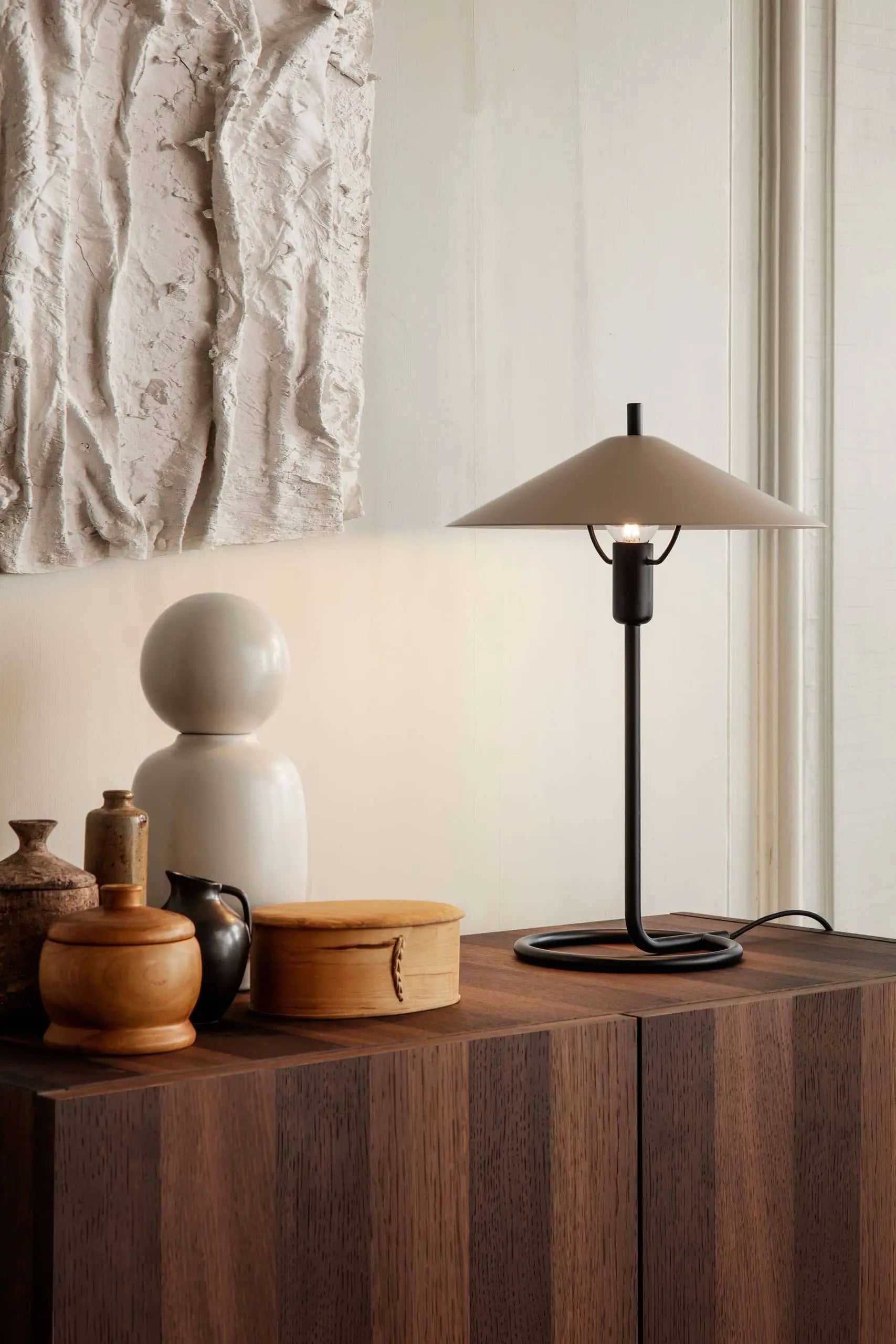 The ferm LIVING Filo Table Lamp, crafted from powder-coated iron with a white conical shade, rests on a dark wooden cabinet surrounded by decorative ceramics and wood accents—an elegant addition to any modern home.