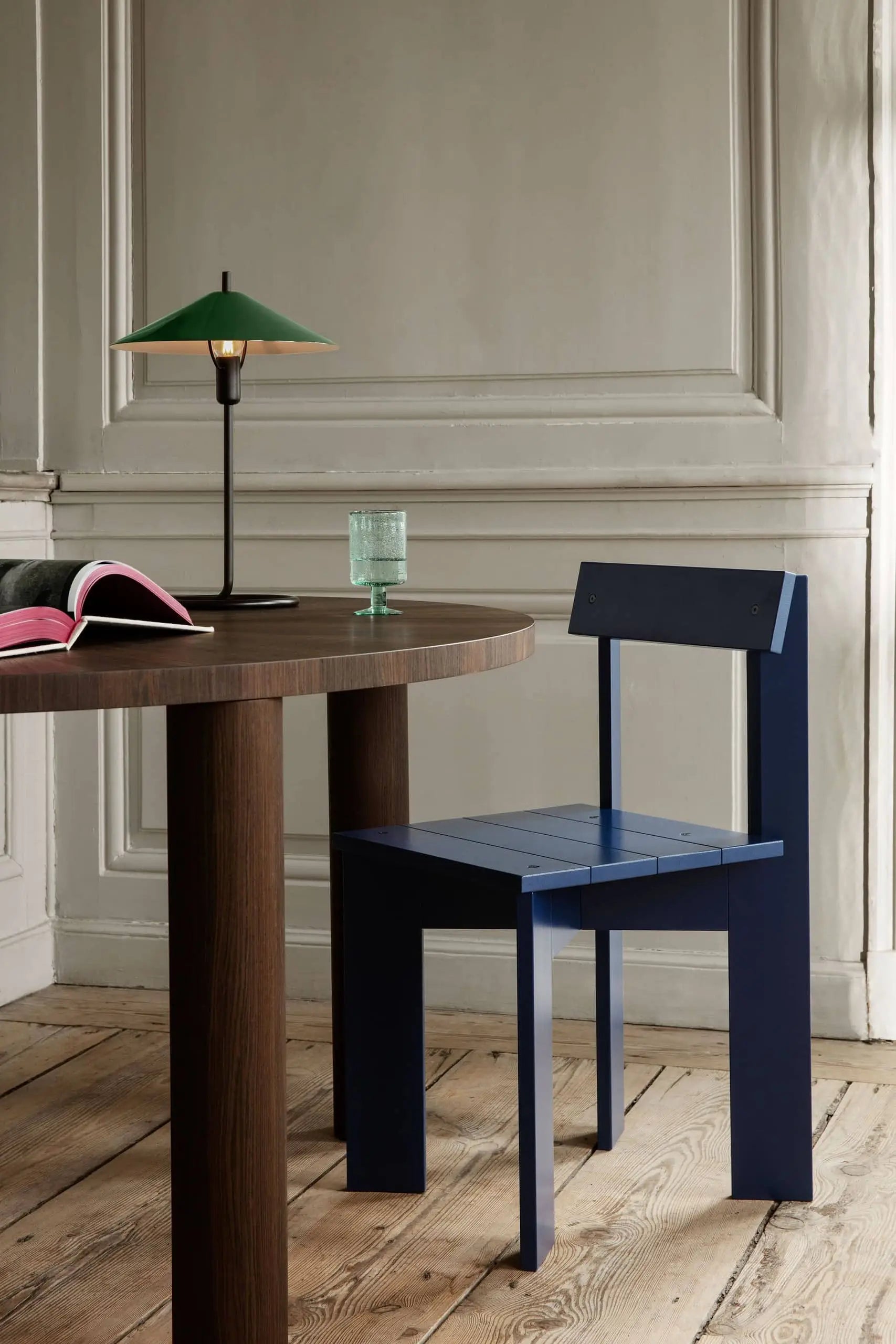 A minimalist room with a round wooden table, modern blue chair, open book, glass of water, and the ferm LIVING Filo Table Lamp in powder coated iron—set against white paneled walls and wood floors for a sleek modern look.