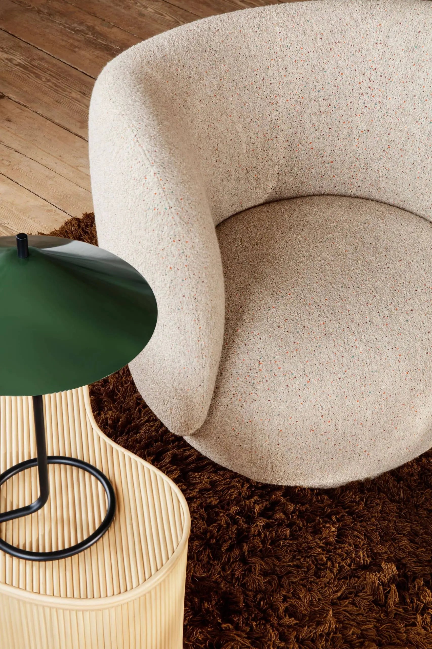 A top-down view of a rounded beige upholstered chair on a brown shaggy rug sits next to the ferm LIVING Filo Table Lamp by ferm LIVING, which rests on a ribbed wood side table, all styled in a cozy modern home.