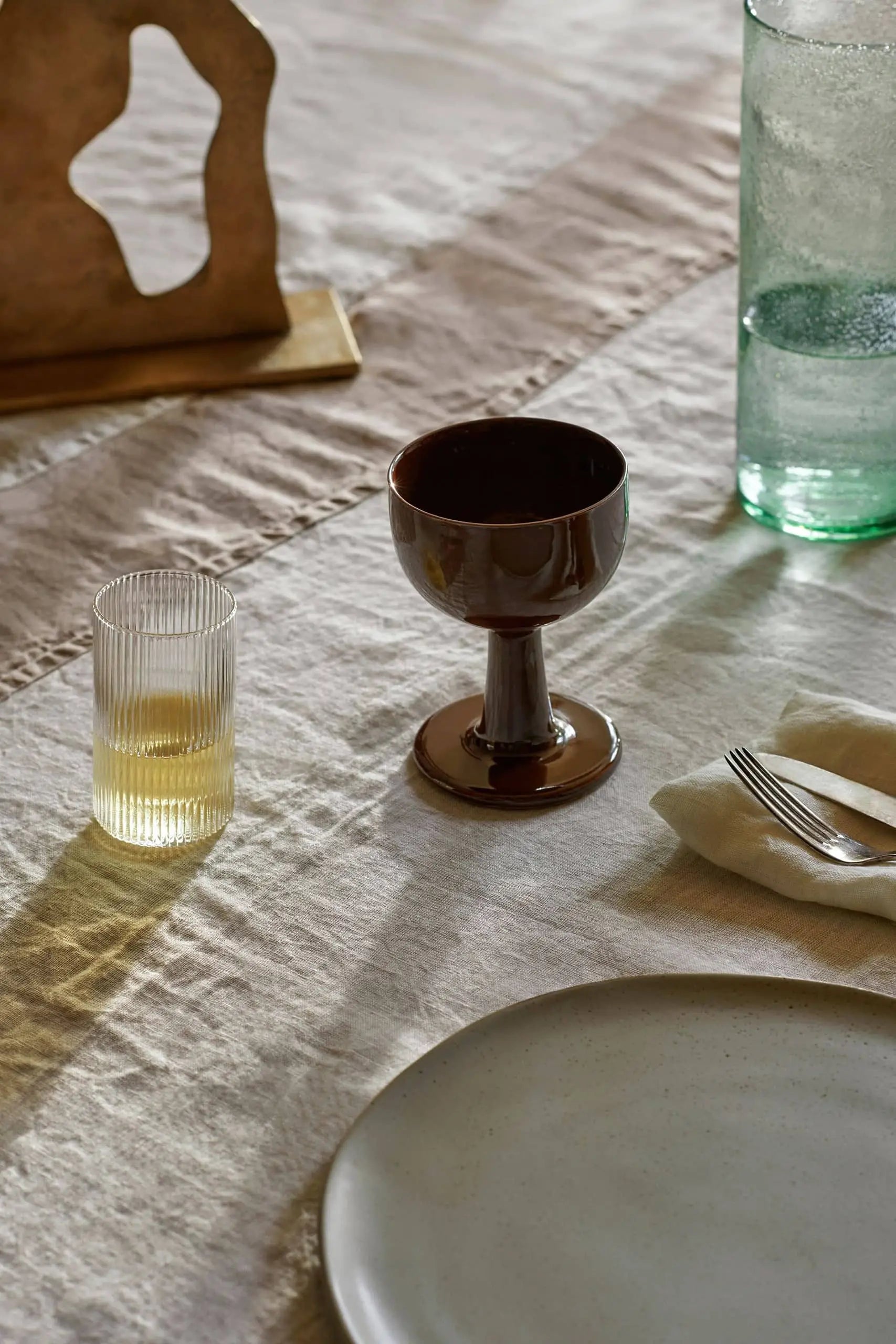 A set table features the ferm LIVING Floccula Wine Glass, a ribbed glass with a yellow drink, green bottle, cutlery on a cloth napkin, and ceramic plate on a beige tablecloth—evoking cozy French bistro charm in warm natural light.