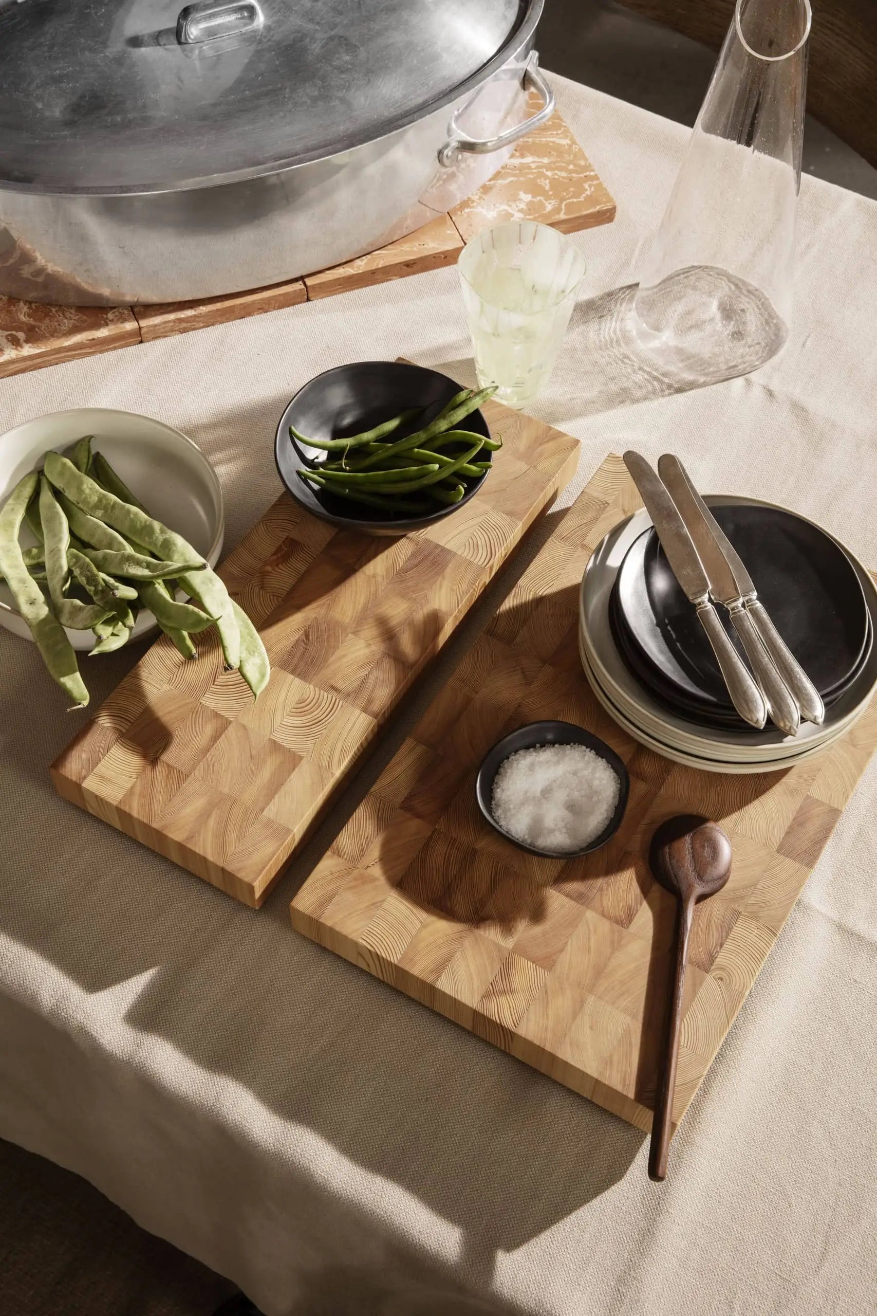 Two wooden cutting boards on a table with green beans, artisanal tableware, plates, knives, a ferm LIVING Flow Porcelain Bowl, Small, a small bowl of salt, glass of water, carafe, and a large pot partially visible in the background.