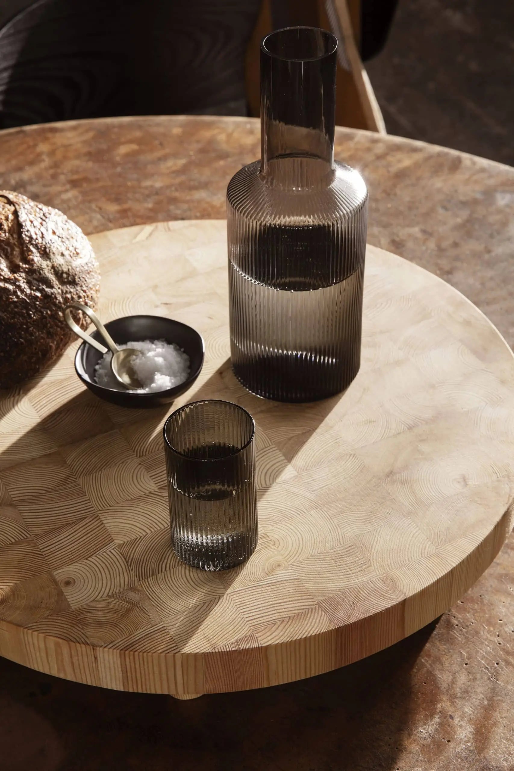 A ribbed glass carafe and matching glass by ferm LIVING stand on a round wooden table. Beside them, artisanal bread and a ferm LIVING Flow Porcelain Bowl, Small, with salt, sit on a wooden board in warm natural light.