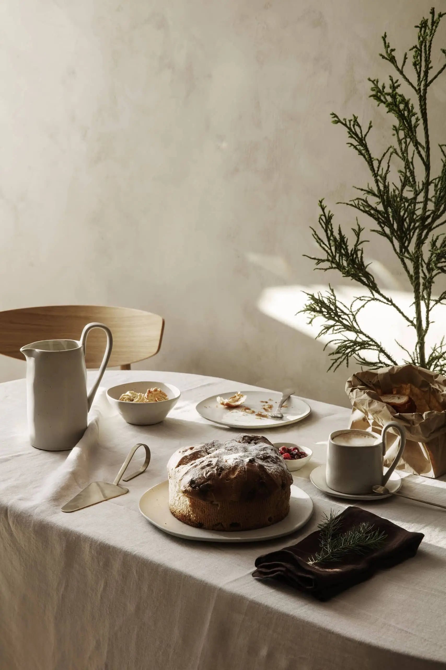 A minimalist table with a white cloth displays a round cake, the ferm LIVING Flow Porcelain Bowl (Small), ceramic pitcher and cup, and greenery in a paper bag. Soft light enhances the calm, cozy atmosphere.