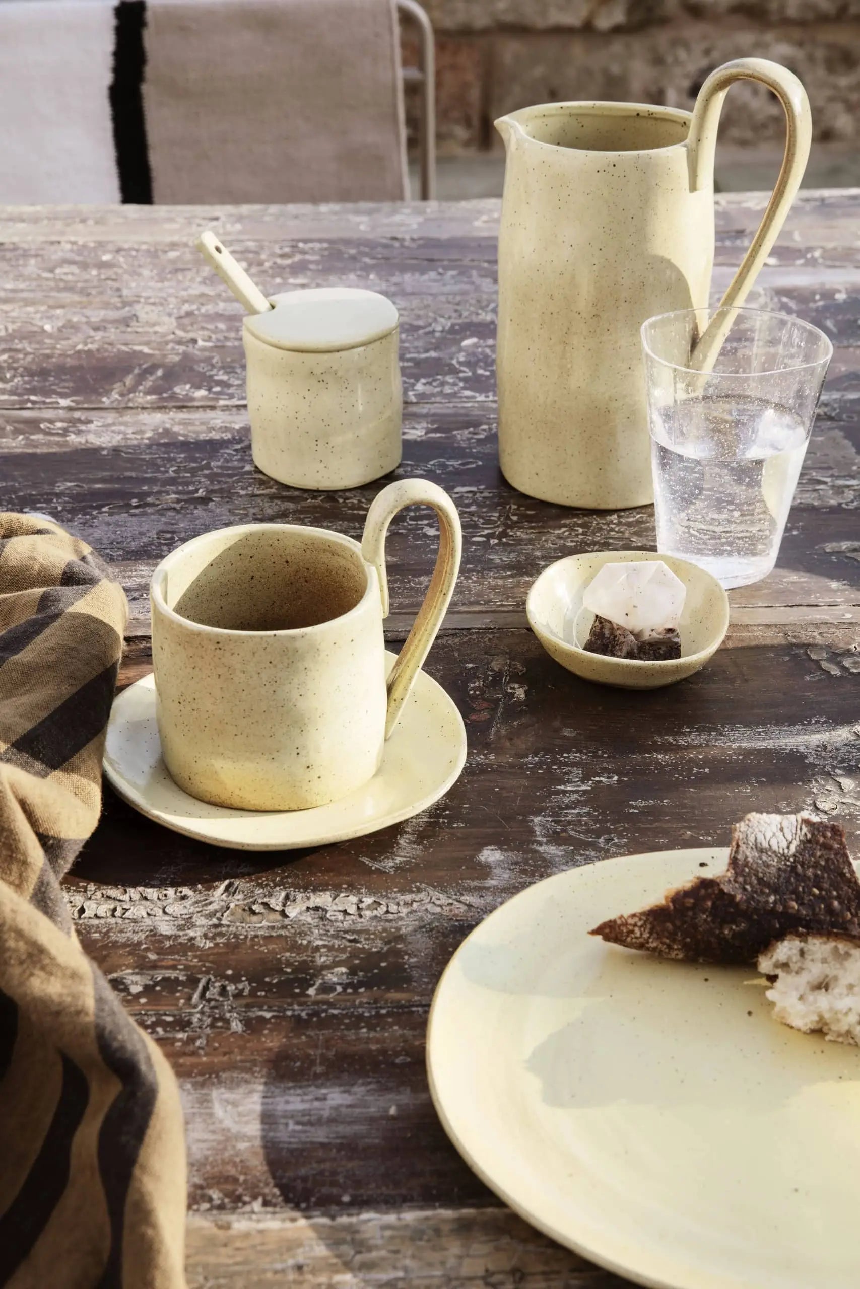 A rustic wooden table set with artisanal tableware, including a beige cup, saucer, jug, sugar jar with spoon, ferm LIVING Flow Porcelain Bowl (Small), glass of water, bread plate, and a striped cloth.