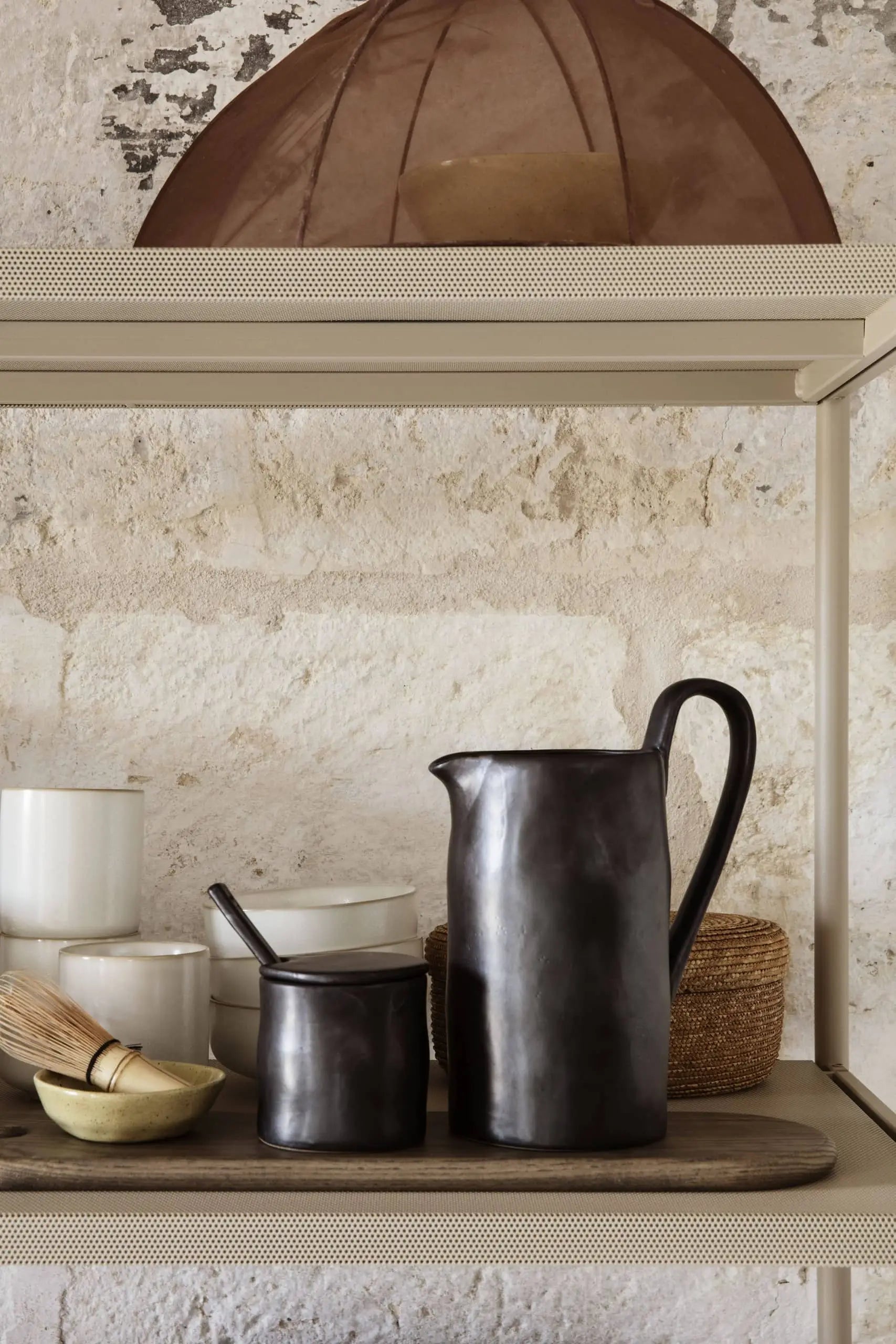 A beige shelf showcases the ferm LIVING Flow Jug, a matching cup, white kitchen containers with lids, a bamboo whisk in a brass bowl, and a woven basket set against a textured stone wall.