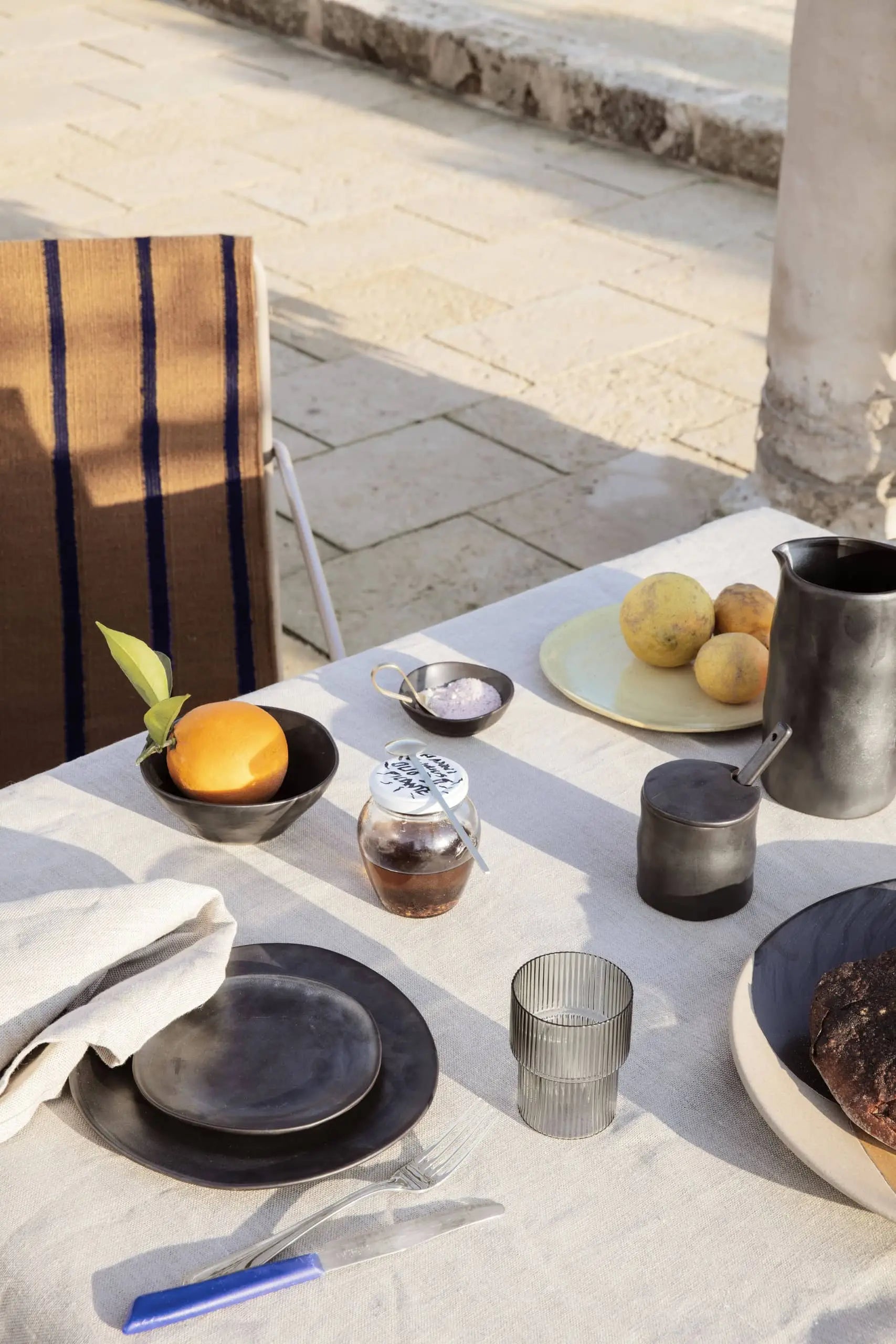 A sunlit outdoor table set with black ferm LIVING Flow Plate, Small by ferm LIVING, a glass, a pitcher, jam jar, orange, lemon, pear, bread loaf, folded napkin. A striped chair is partially visible next to the table.