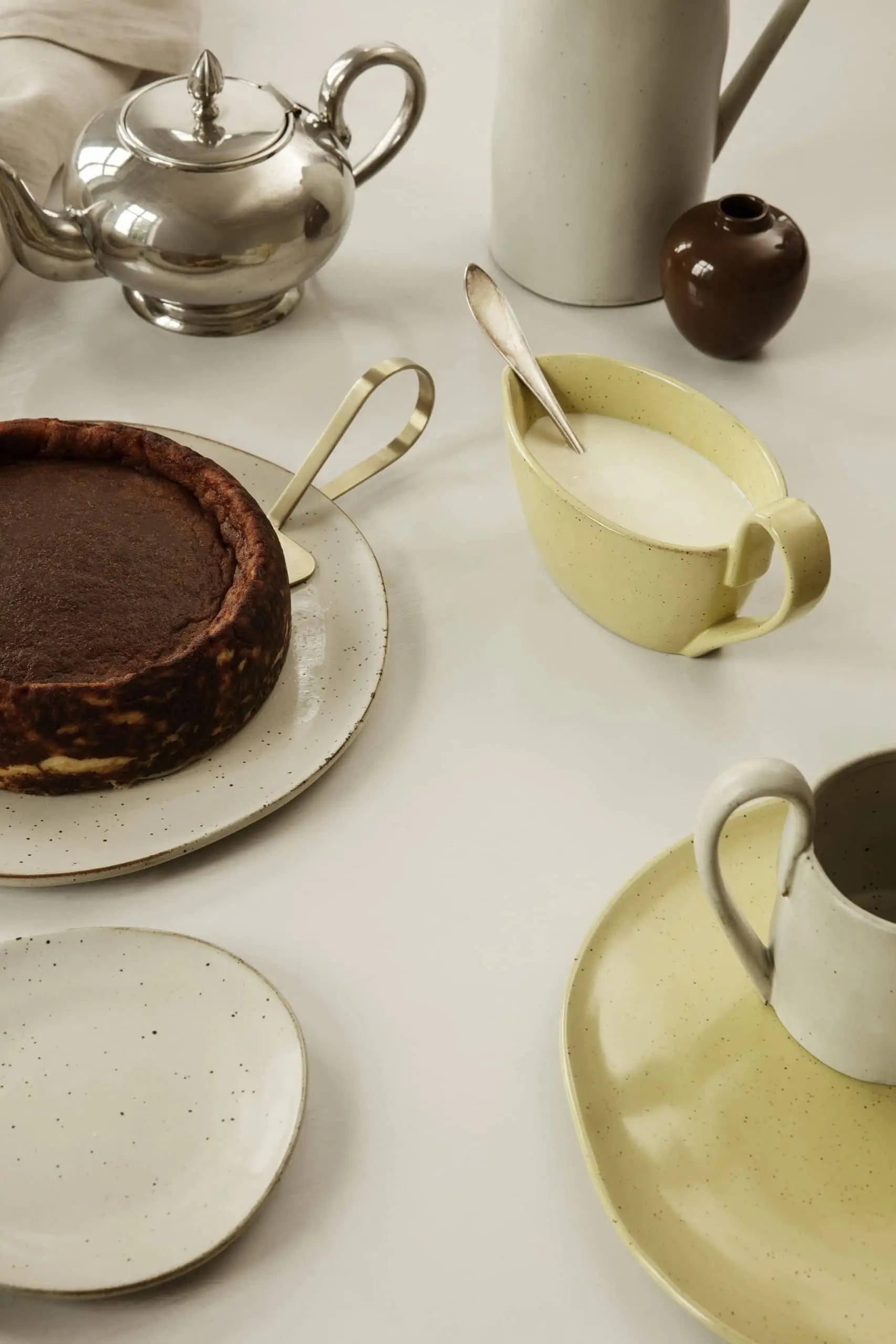A table set with chocolate cake, coffee, milk, plates, and a teapot in neutral tones features the ferm LIVING Flow Plate, Small by ferm LIVING—crafted from porcelain with a subtle reactive glaze for an elegant look.