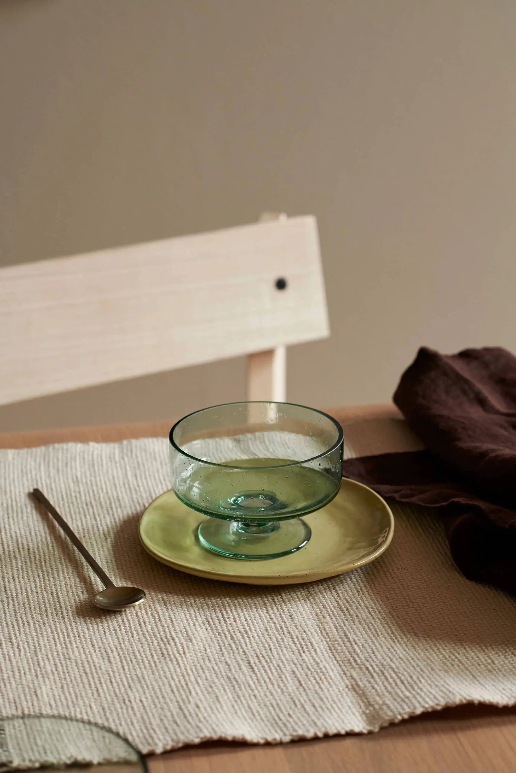 A clear green glass dessert bowl rests on a small ferm LIVING Flow Plate in yellow, placed on a beige placemat beside a silver spoon and dark brown napkin, with a light wooden chair behind.