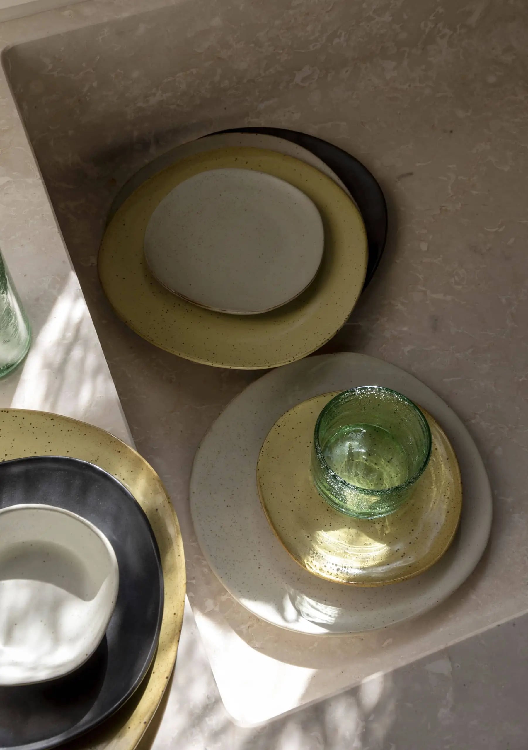 A ferm LIVING Flow Plate, Small, in a neutral hue and a green glass cup sit on a beige countertop, sunlight casting soft shadows across the surface.