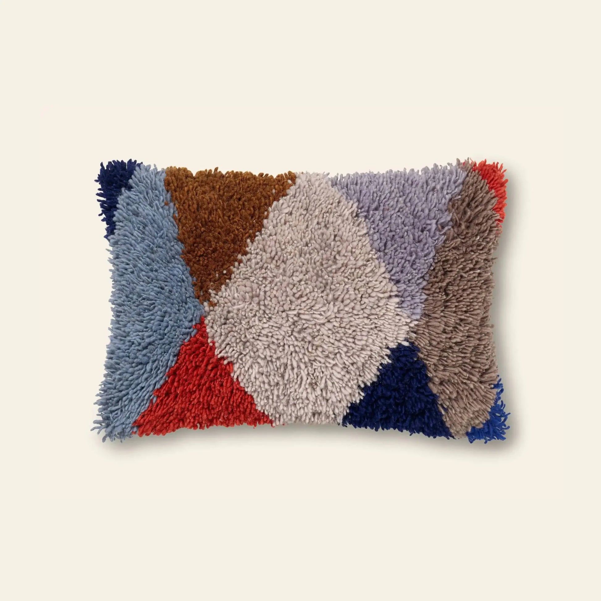 The ferm LIVING Harlequin Tufted Cushion is a rectangular pillow with a shaggy, textured surface and geometric shapes in blue, brown, red, gray, beige, and tan on a light background.