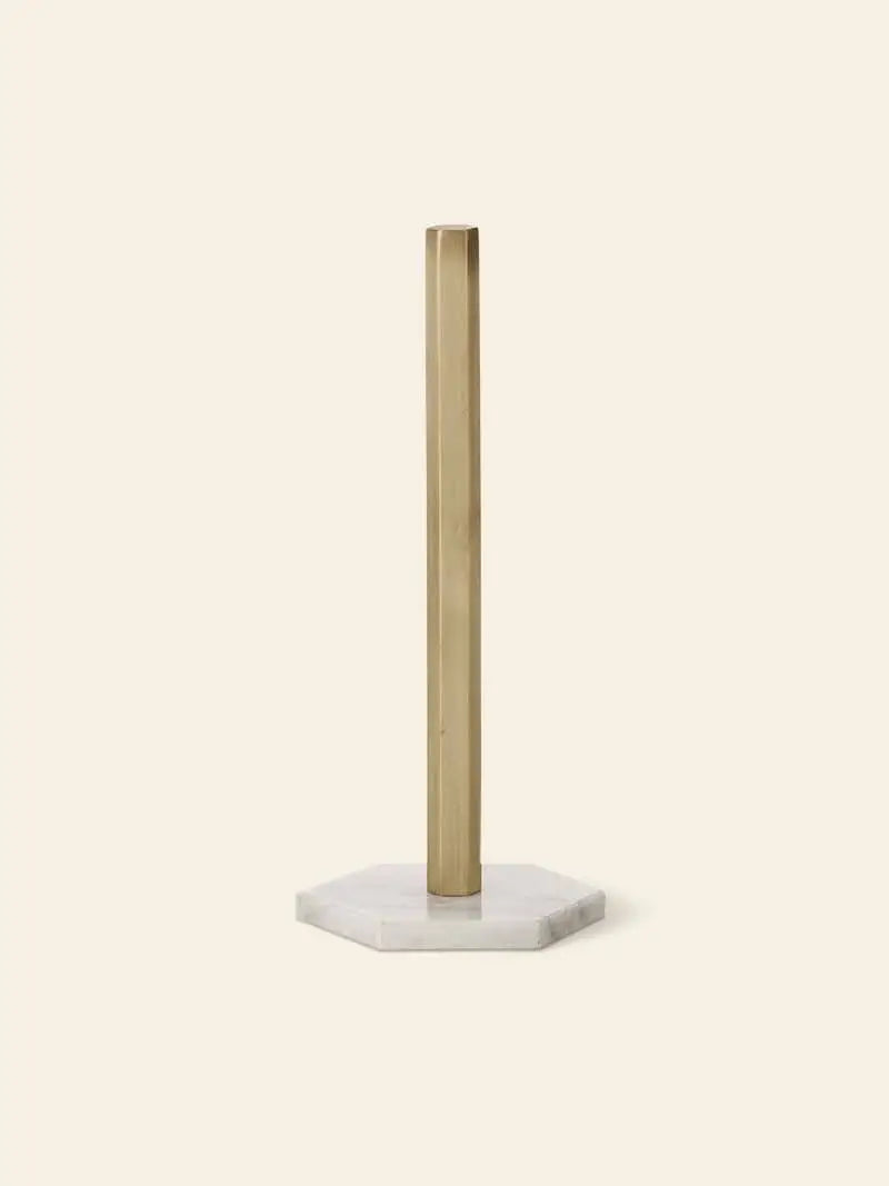 The ferm LIVING Hexagon Stand is a minimalist paper towel holder with a brass-finished vertical rod and white marble hexagon base, designed by ferm LIVING to hold rolls neatly on a light beige background.