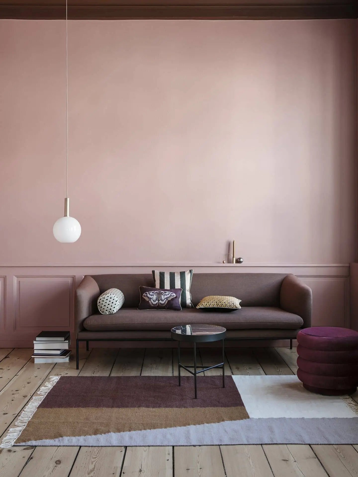 A modern living room with a mauve sofa, patterned cushions, a round purple ottoman, two geometric glass coffee tables, books, a white globe pendant light, a muted pink paneled wall, and the ferm LIVING Kelim Rug by ferm LIVING.