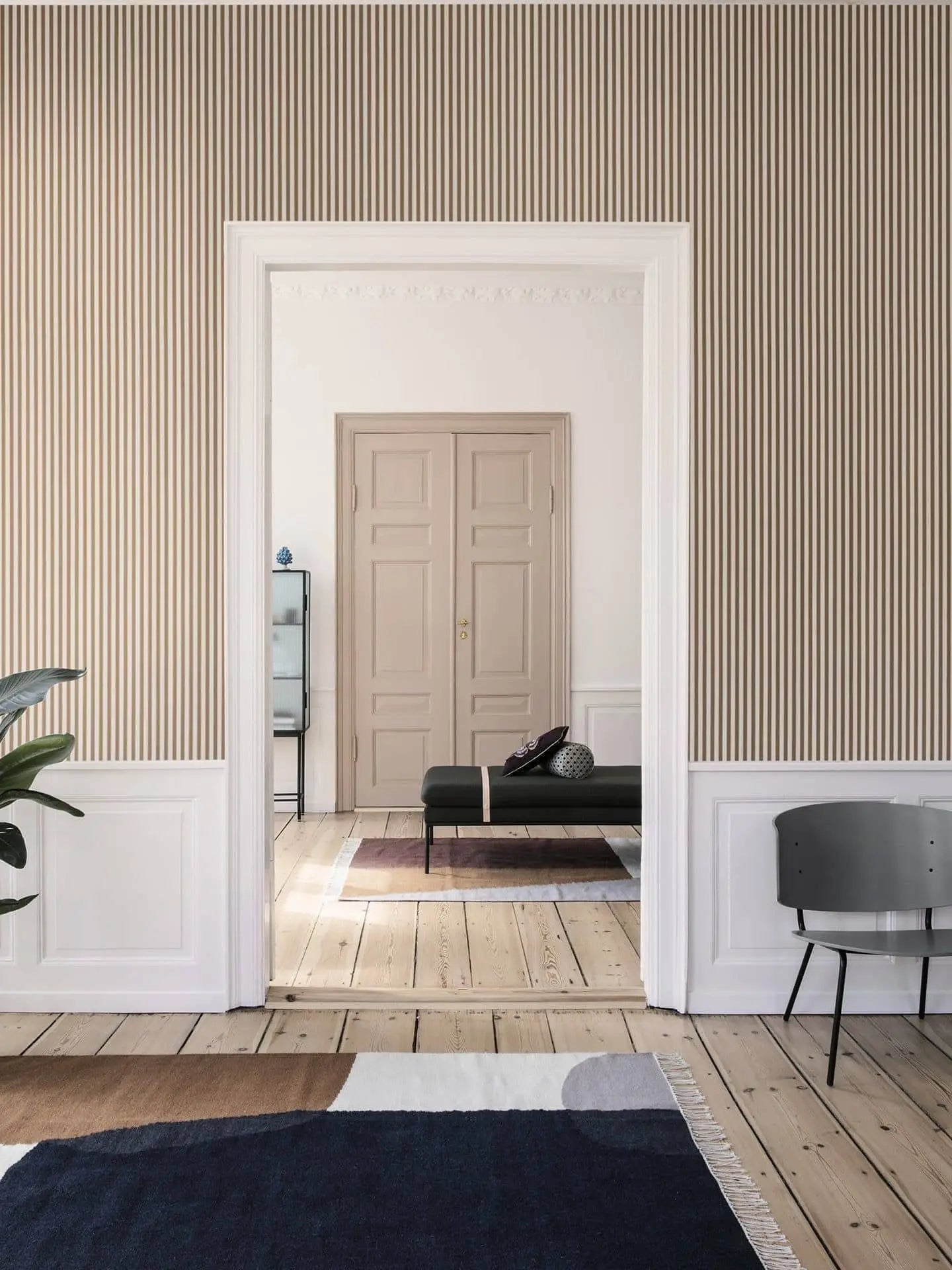 A minimal, modern space with beige vertical striped wallpaper, wood floorboards, a black bench, ferm LIVING Kelim Rug with geometric shapes, two-door closet, and simple decor including a gray chair and potted plant.