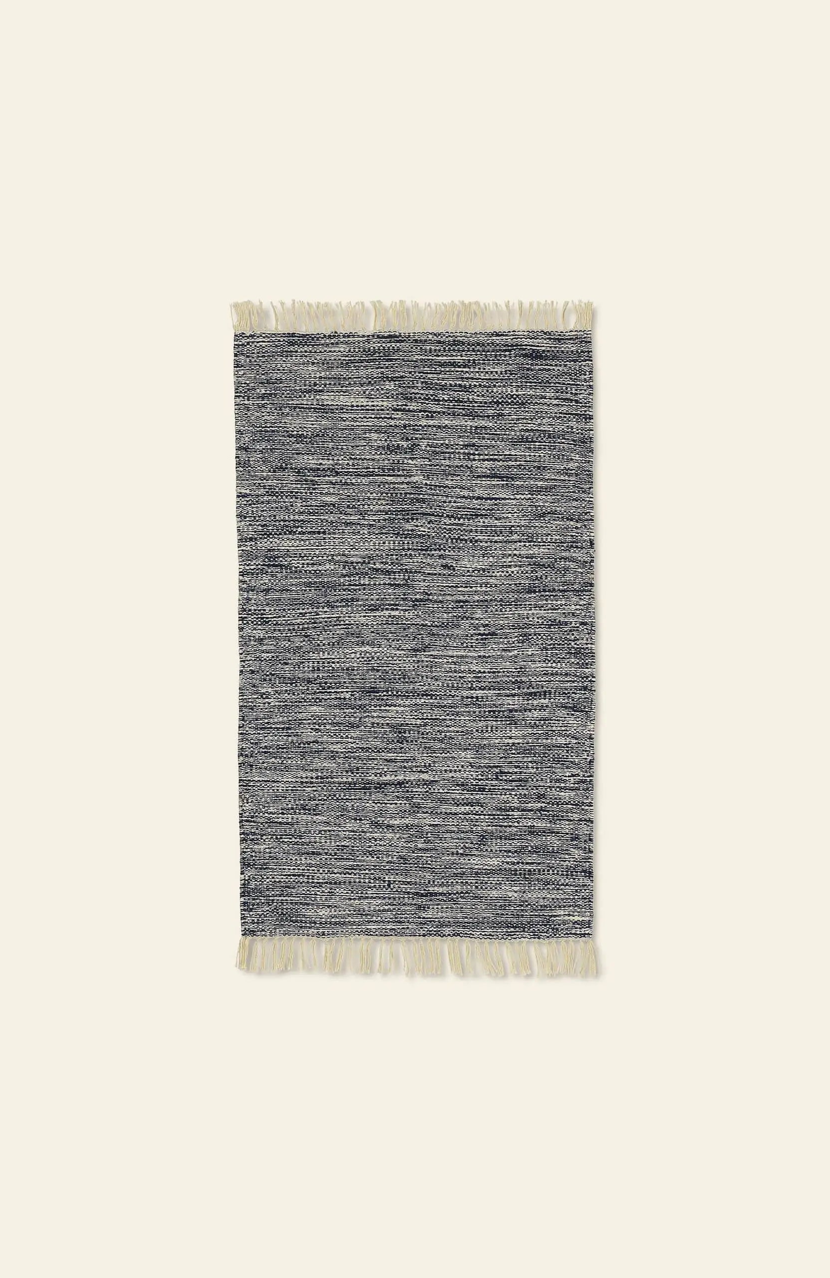 The ferm LIVING Melange Rug by ferm LIVING features a rectangular shape in gray and white with a textured pattern and short cream fringe on the ends, providing an easy-care, luxurious look, shown against a plain light background.