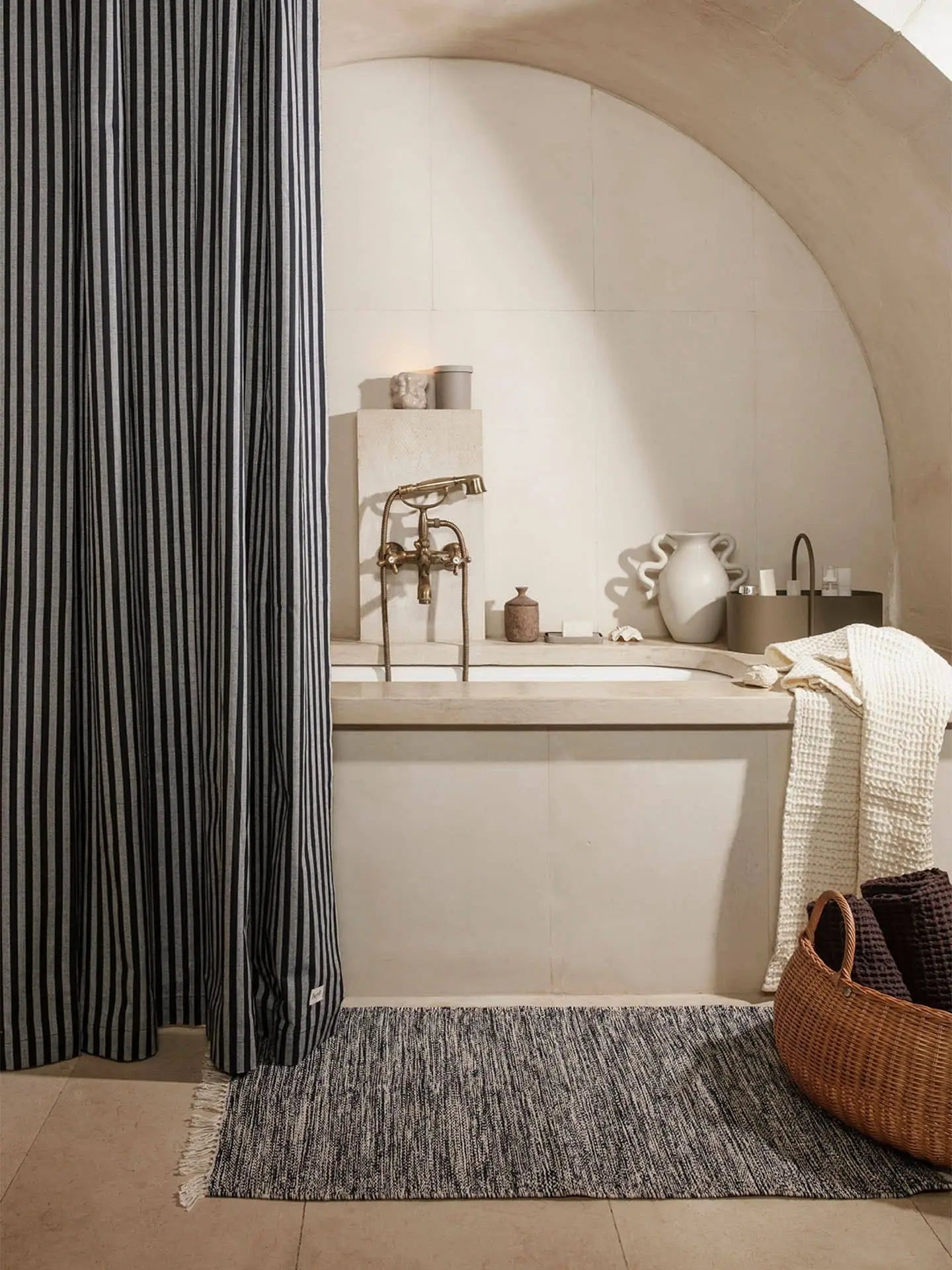 A modern bathroom with a built-in tub, arched ceiling, and neutral tiles features a striped shower curtain, ferm LIVING Melange Rug, woven basket, and ceramic decor for added warmth and minimalist style.
