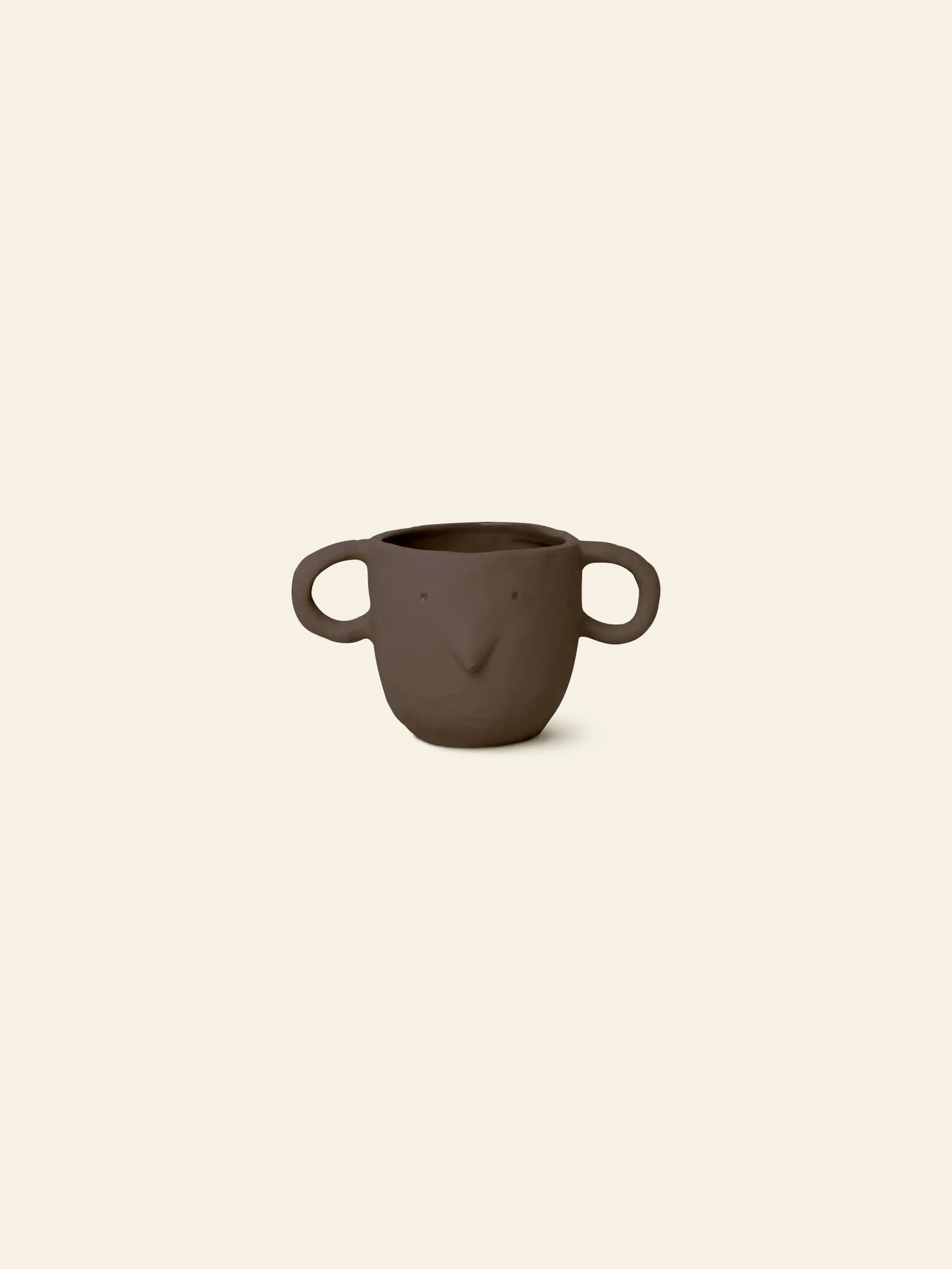 The ferm LIVING Mus Plant Pot, Small features two large mouse handles and a simple face with dot eyes and a pointed nose, crafted in brown ceramic and inspired by the playful style of ferm LIVING. Set against a plain beige background.