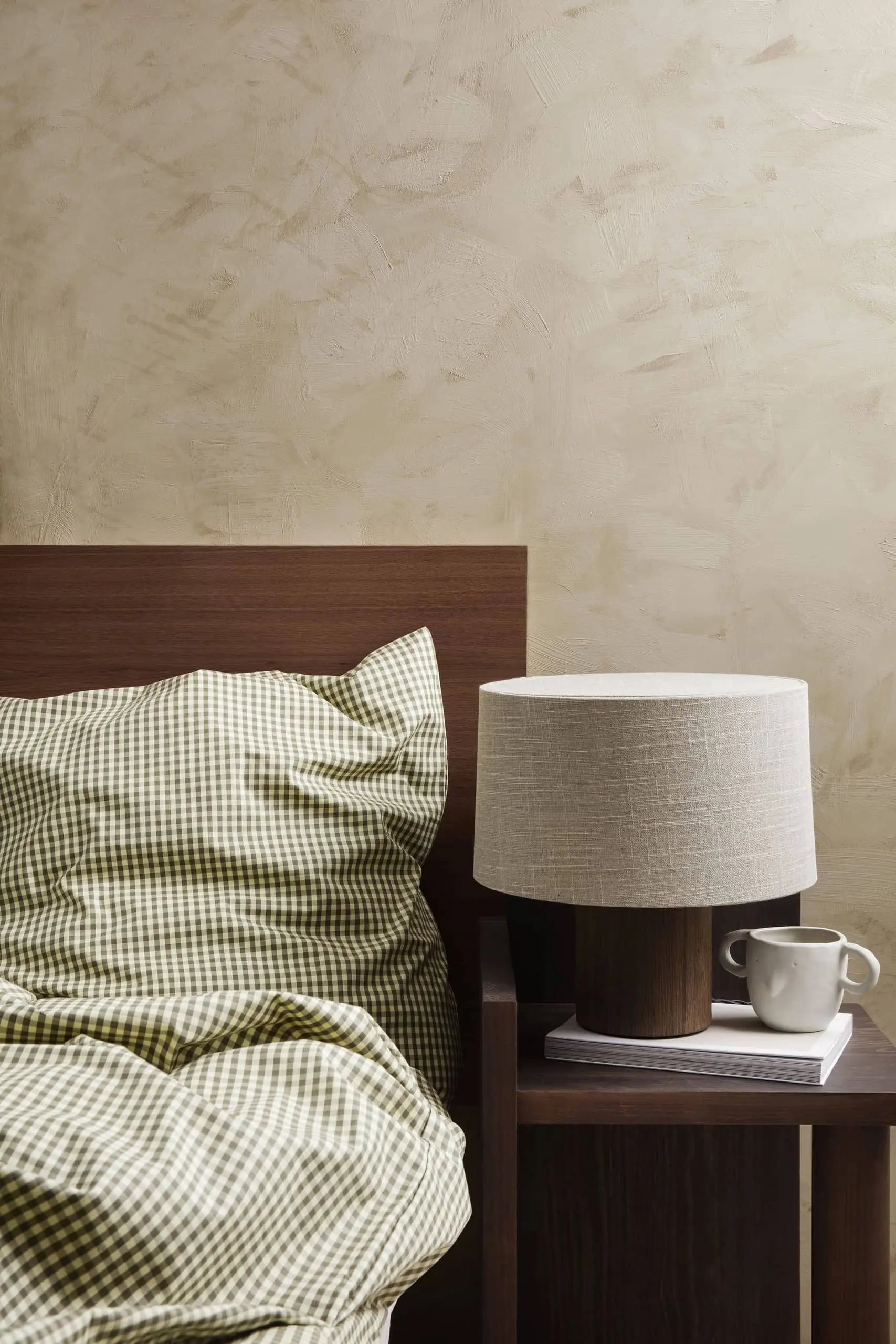A neatly made bed with green and white checkered bedding is next to a wooden nightstand holding a ferm LIVING Mus Plant Pot, Small, a table lamp, a white cup, and two books against a beige textured wall.