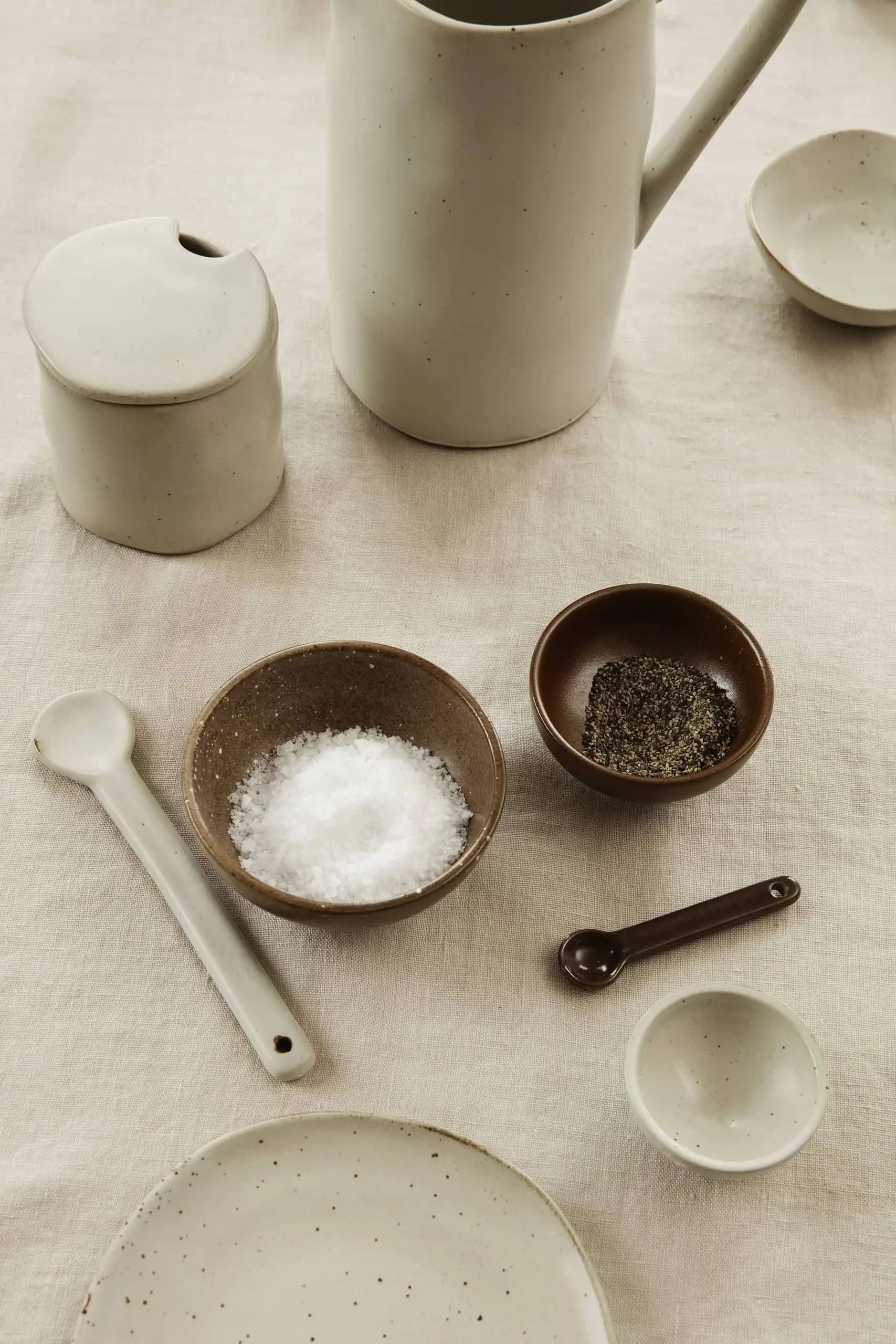 A minimalist table setting with ceramic dishes and utensils, featuring a jug, lidded container, ferm LIVING Petite Bowls Set of 3 for salt and pepper, two measuring spoons, and small dishes—all atop a neutral cloth for an elevated dining experience.