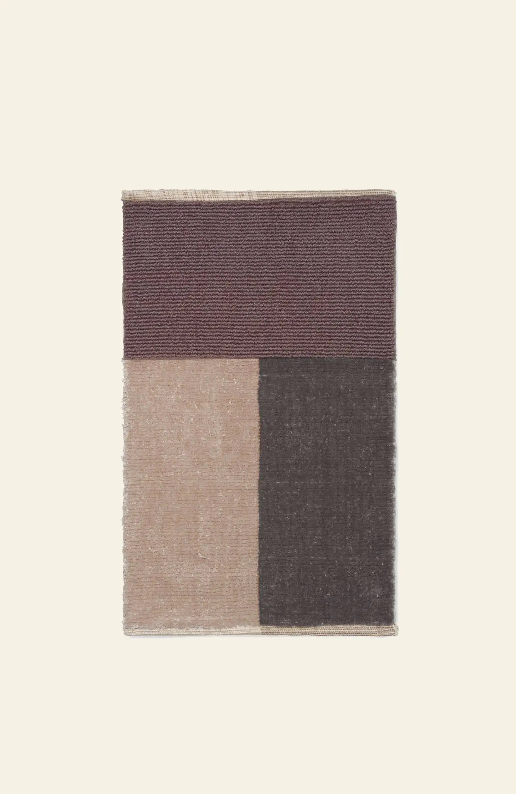 The ferm LIVING Pile Bathroom Mat features ribbed dark maroon, smooth beige (left), and dark brown (right) color blocks. Made from recycled polyester for a luxurious feel, it’s set against a light beige background.