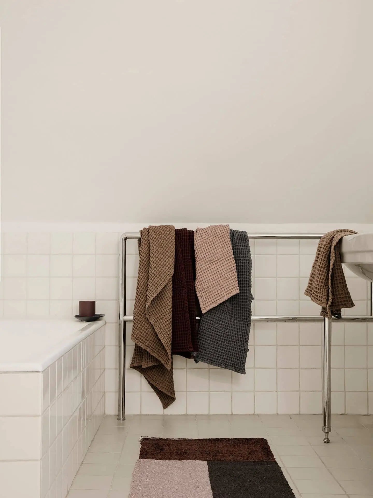 Minimalist bathroom with white tiles, a metal rack with brown, pink, and dark gray towels, a candle on the tub edge, and the ferm LIVING Pile Bathroom Mat—crafted from recycled polyester for a luxurious underfoot feel.