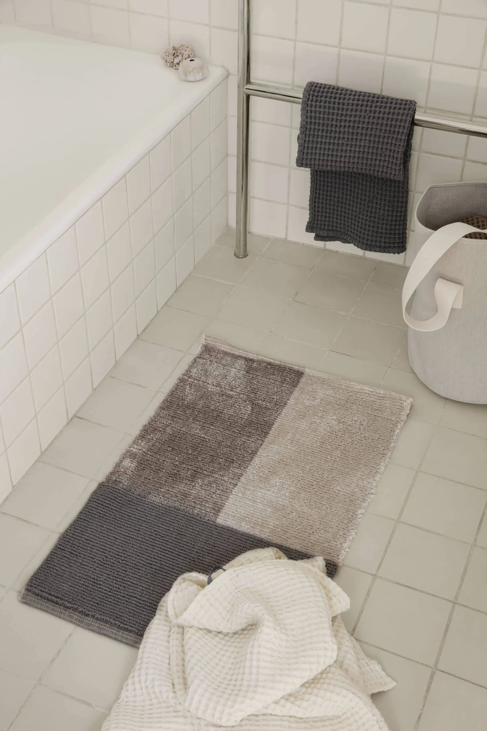 A modern bathroom with white tile features the ferm LIVING Pile Bathroom Mat in taupe and gray, made from recycled polyester for a luxurious touch. Folded towels rest on a metal rack, with a gray handled laundry basket nearby.