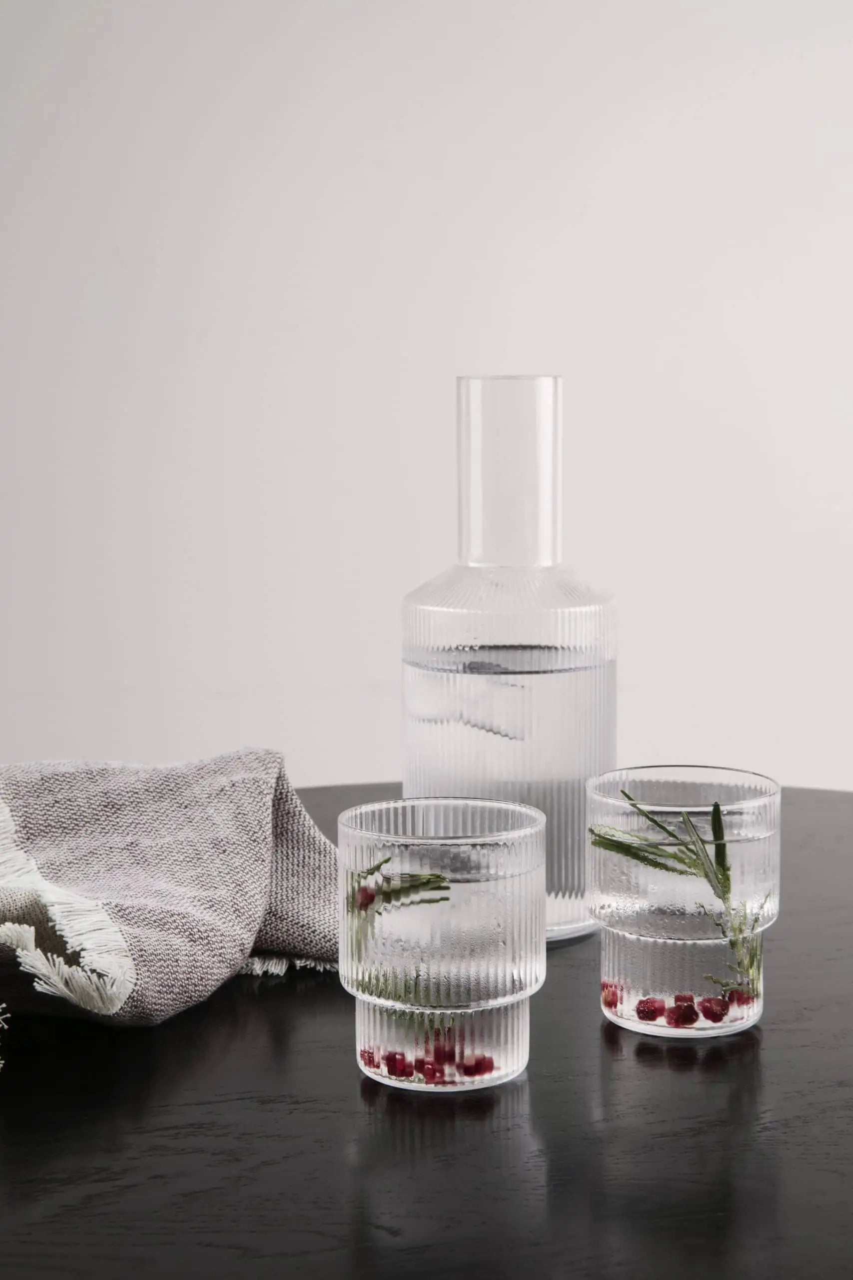 A glass carafe and two ferm LIVING Ripple Drinking Glasses, filled with water, rosemary sprigs, and pomegranate seeds, sit on a dark table beside a folded gray cloth—a stylish set from ferm LIVING.