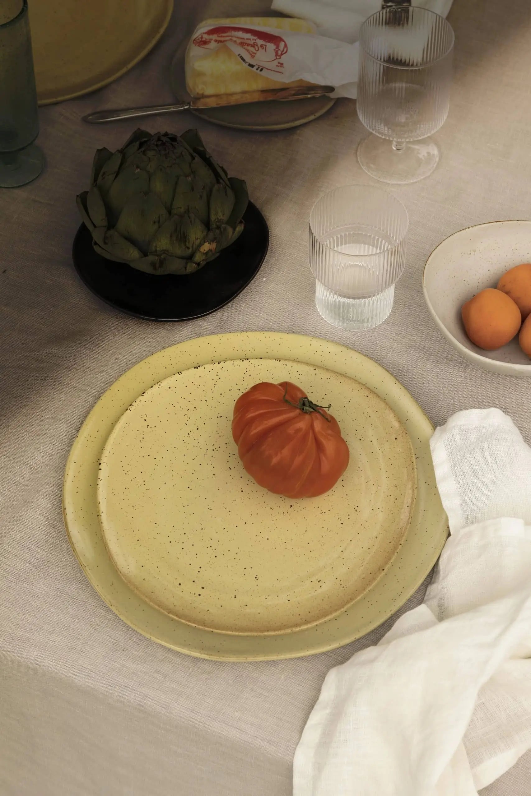 A speckled beige plate with an heirloom tomato sits on linen, surrounded by a napkin, artichoke, eggs in a bowl, and a set of ferm LIVING Ripple Drinking Glasses completes the scene.